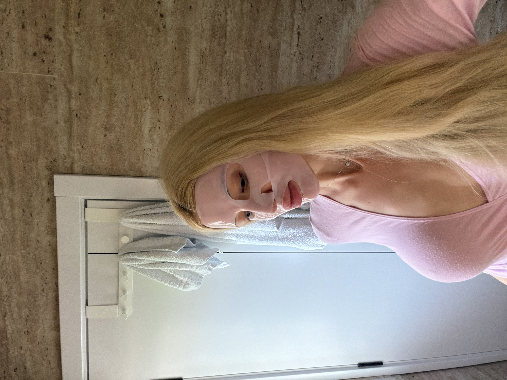 A woman with long blonde hair and a pink face mask is wearing a light pink shirt. She is in a bathroom with beige tiled walls and a white cabinet. A white towel is hanging on the cabinet door. It is not possible to ascertain the location in Belgrade, Serbia.