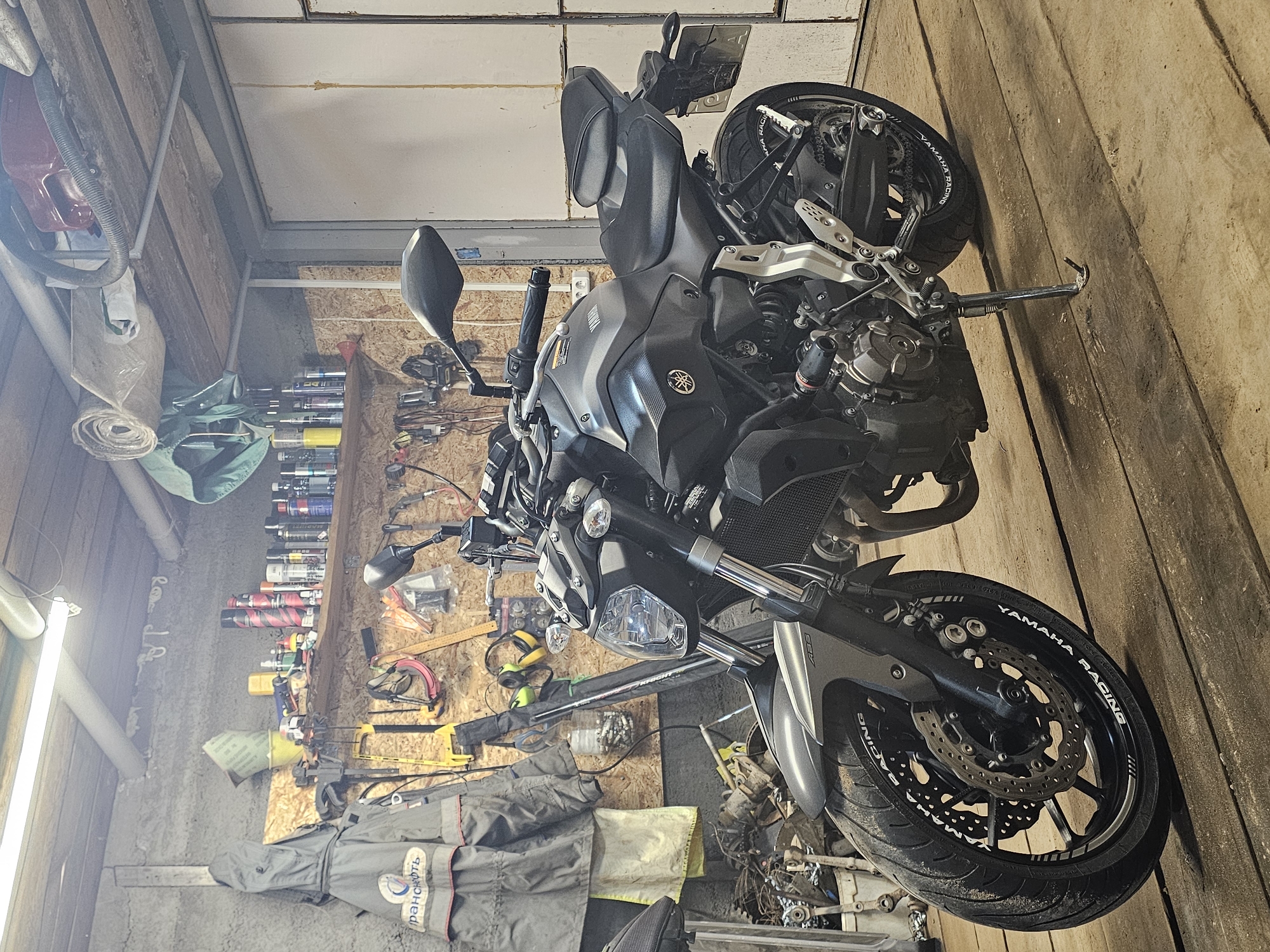 The image shows a gray Yamaha MT-07 motorcycle parked inside a garage. The garage walls are made of wood and insulated with styrofoam. There's a workbench with various tools, spray paint cans, and other objects mounted on the wall. A gray coverall lies on the floor. Based on information on the coverall, the location is Уральское, Russia.