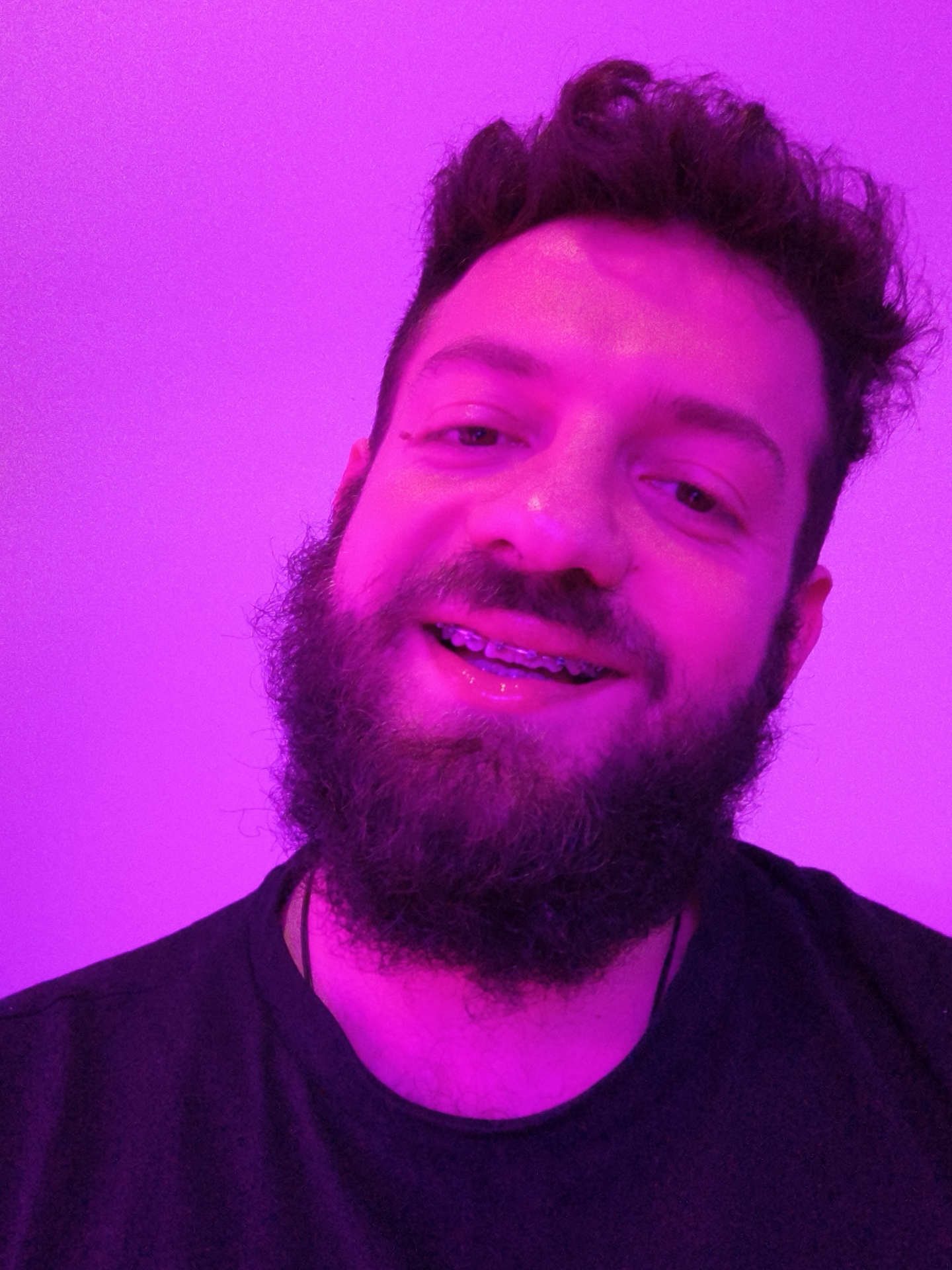 A selfie of a man with dark hair and a beard, wearing braces. The image is lit with a purple light, making it difficult to discern finer details. The man is wearing a black t-shirt. The background is a plain wall, also illuminated by the purple light. There is no evidence to suggest the location is Diepholz, Germany.