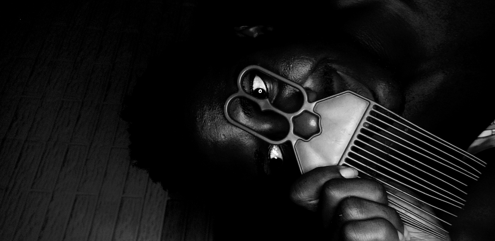 The image is a black and white close-up of a person holding a hair comb. The person's face is partially visible, with one eye peeking through one of the comb's handle holes. The comb is held in the person's hand, with the teeth of the comb extending outwards. The image is dark, with the person's skin and hair appearing dark against the background. There is a wall partially visible on the left side. The location is unspecified beyond the city, with no landmarks visible in this scene.