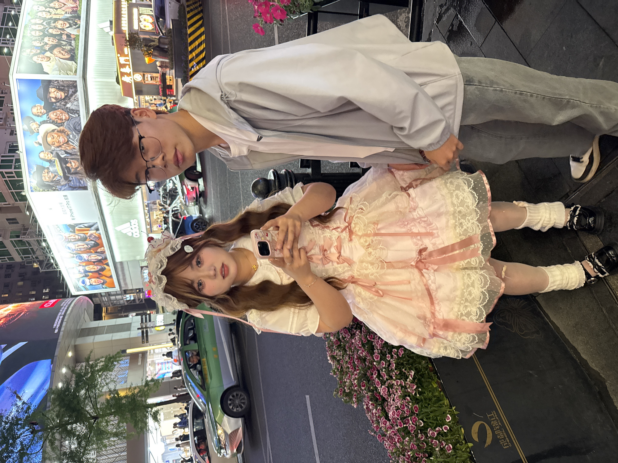 A young adult couple is photographed on a street in Jinjiang District, China, at night. The woman wears a light pink, frilly dress with white knee-high socks and black shoes. She is holding a smartphone, presumably taking a photo. The man, wearing eyeglasses, a light grey hoodie and grey jeans, stands behind her. The street scene includes traffic with green and white taxis, buildings with advertisements, and a flower bed.