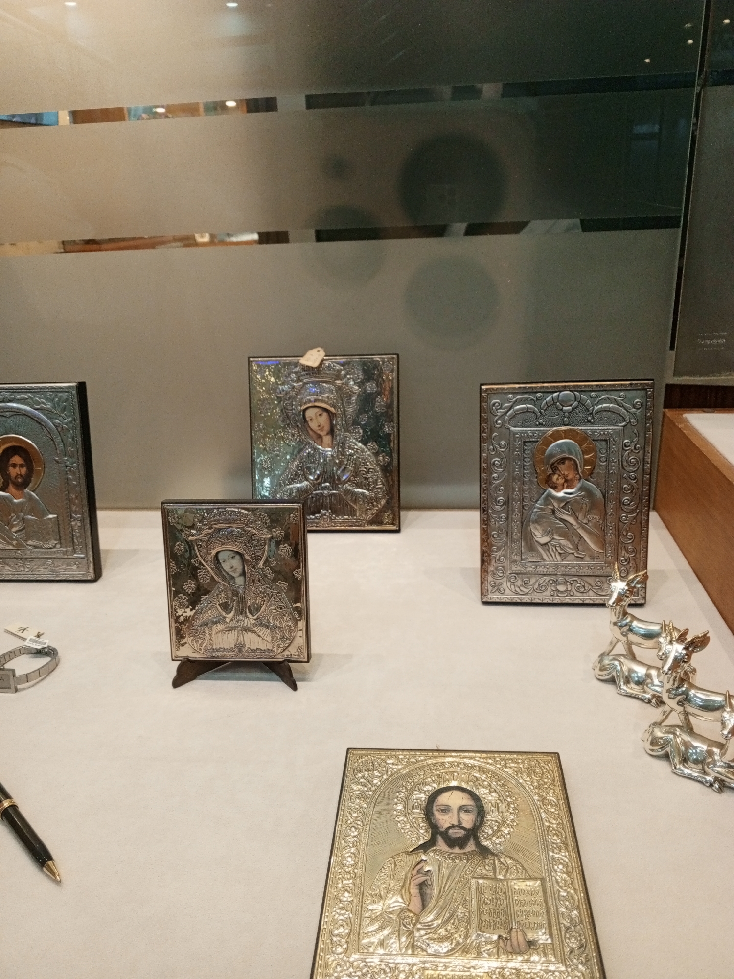 The image shows a collection of religious icons displayed on a white surface. The icons appear to be silver or metal-plated, depicting various Christian figures including Jesus Christ and the Virgin Mary. There are five visible icons in different sizes and designs, including one resting on a small easel. The display also includes a pen, a watch, and a set of silver figurines, possibly deer. The background suggests a shop or exhibit space, with a frosted glass panel and a wooden shelf visible. The image was taken in Porlamar, Venezuela.