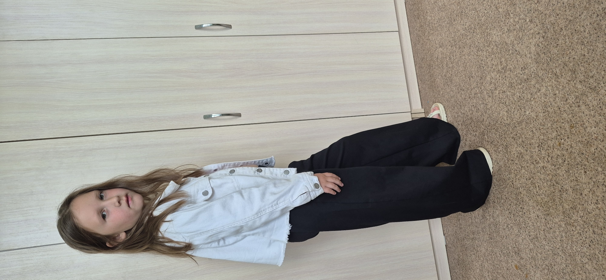 A young girl is sitting on the floor in an indoor environment. She has light brown hair and is wearing a white denim jacket and black pants. Her feet are shod, with one foot appearing to be in a white sandal. She is seated next to a light-colored dresser with metal drawer pulls. The floor is covered with a brown carpet. The location appears to be a room in a home or apartment in an unknown city.