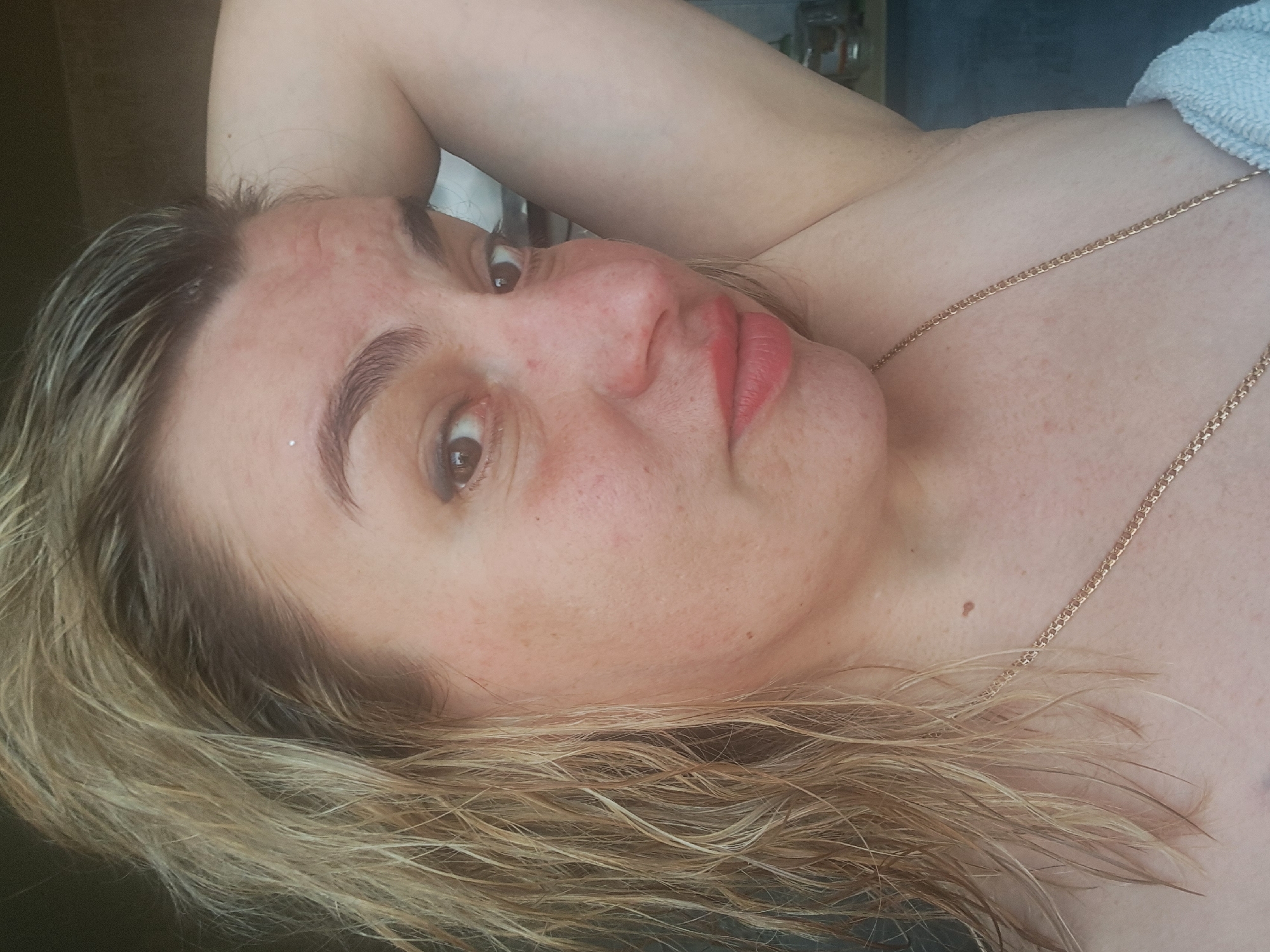 A medium shot, rotated counter-clockwise, shows a middle-aged Caucasian woman with blonde hair. Her hair appears wet. The woman is reclining, with one arm raised behind her head. She is wearing a thin gold chain necklace. The background includes a blurred view of what appears to be a bathroom, with toiletries visible on a shelf. There is no information to determine the location of the photo in Elektrostal, Russia.