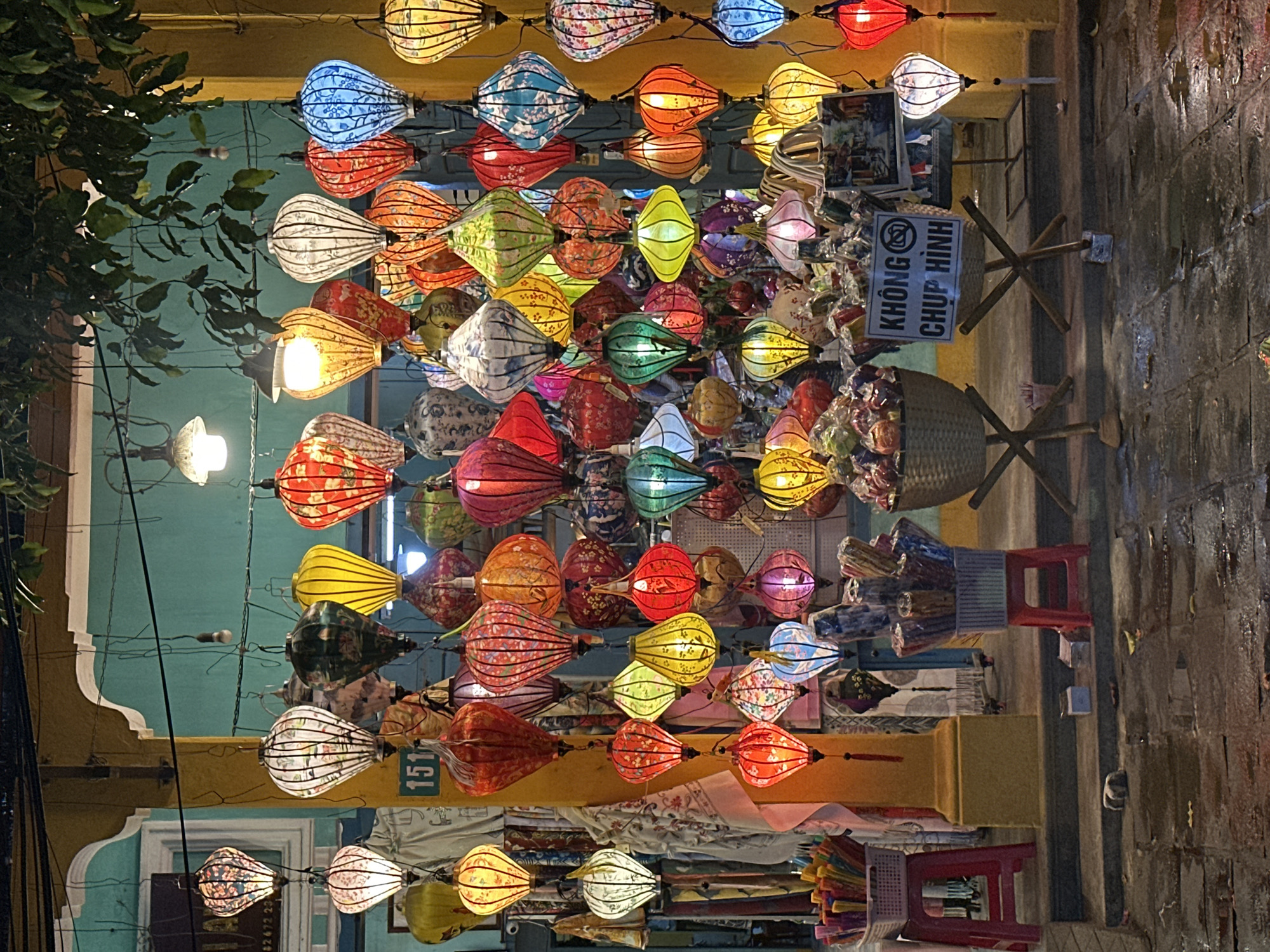 This is a vertical, eye-level shot of a street market in Hoi An, Vietnam, at night. The scene is dominated by numerous hanging lanterns of various colors and patterns, illuminated and creating a vibrant, festive atmosphere. The lanterns are strung together in rows, filling the frame from top to bottom.
Among the lanterns, some are solid colors like red, orange, yellow, blue, and green, while others feature intricate floral or geometric patterns. Some are lit from within, casting a warm glow, while others appear to be unlit.
To the left of the frame, dark green foliage hangs from a tree, adding a natural element to the man-made display. In the background, building facades with yellowish and turquoise paint are visible, characteristic of Hoi An's architecture.
On the right side of the image, a woven basket rests on a wooden stand. A sign with Vietnamese text is attached to the stand. The text reads "KHÔNG CHUP HINH" which translates to "No photography." Red plastic stools and stacks of colorful folded fabrics are also visible, suggesting a retail setting.
The ground appears wet, indicating it might have rained recently or that the street is being cleaned. The overall impression is one of a lively and colorful night market, likely a tourist destination. The time of day is clearly night due to the artificial illumination of the lanterns.