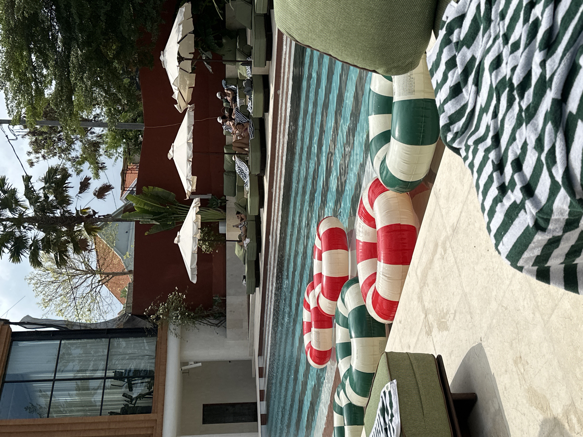 The image depicts a resort-style swimming pool area. In the foreground, several inflatable rings, some red and white, others green and white, float on the water. To the left, a green upholstered bench with a striped towel is visible. Behind the pool, lounge chairs with people are arranged under white umbrellas. The pool itself has a striped pattern on its bottom. In the background, a modern building with large windows and lush greenery can be seen. The sky is overcast. The scene appears to be set in a tropical or subtropical location.