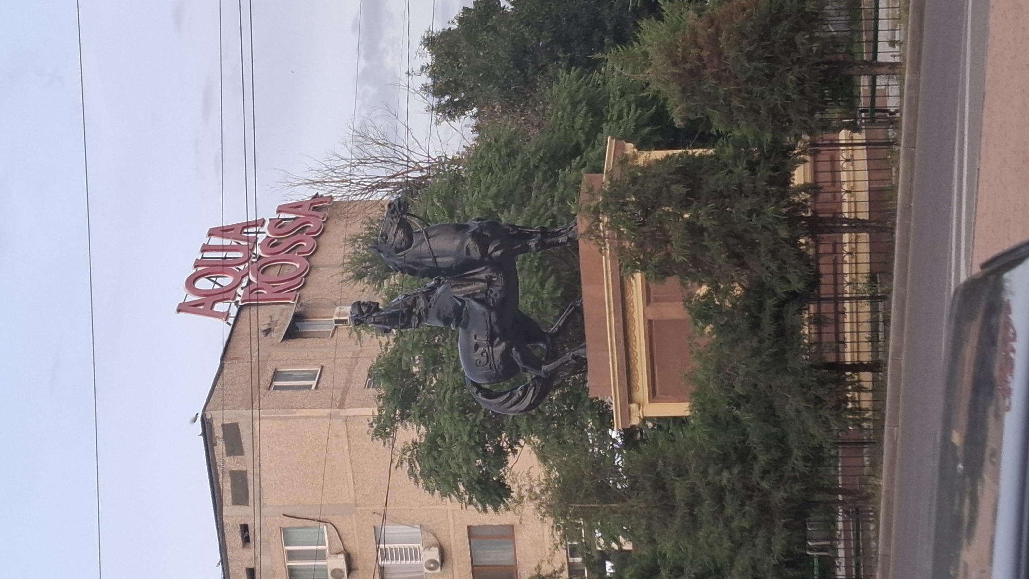 An equestrian statue of a historical figure is prominently displayed in the foreground, facing right. The statue is made of dark material, likely bronze, and depicts a person in traditional attire riding a horse. The statue stands on a multi-tiered pedestal, which is adorned with decorative elements. Behind the statue, a light brown, multi-story building with numerous windows and air conditioning units is visible. The building has a red neon sign on its roof that reads "AQUA ROSSA". The scene is framed by lush green trees and foliage. A metal fence runs along the front of the statue's base, separating it from a paved area and a road in the immediate foreground. The sky is overcast. The image appears to have been taken in Shymkent, Kazakhstan, possibly on a street or public square, given the presence of the statue, building, and road.