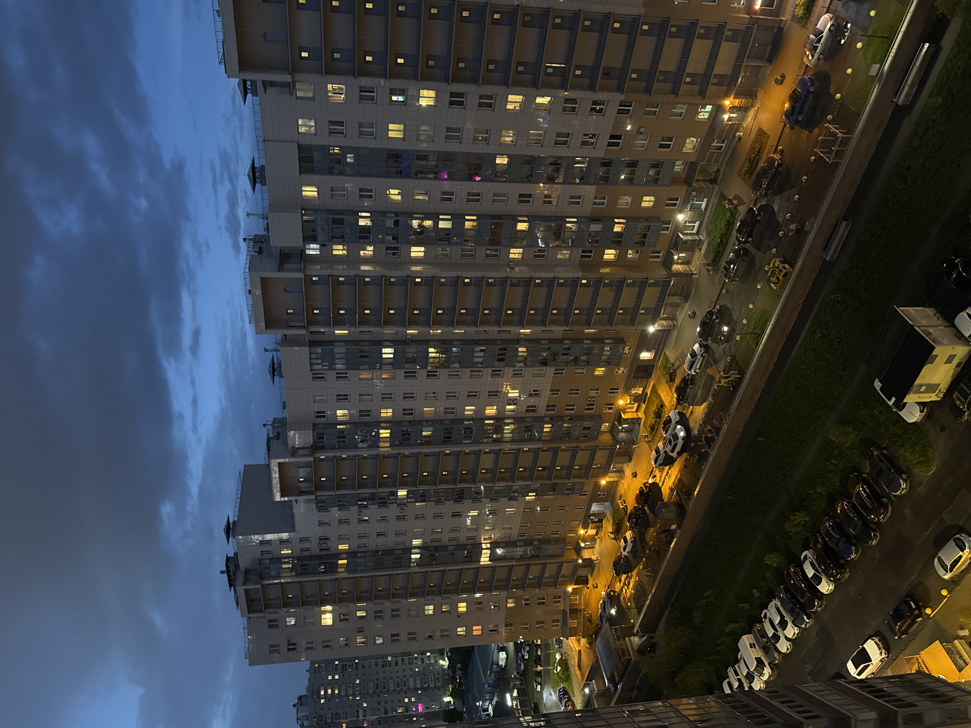 The image shows an aerial view of Sinyavino, Russia at dusk. A large, high-rise apartment building dominates the frame, with numerous illuminated windows. The sky is cloudy and dark. Below the apartment building, a street is lined with parked cars, and a narrow strip of grass separates the cars from the building. Streetlights provide illumination along the street. Another building of similar style is visible in the distance to the left. No people are visibly present in the image.