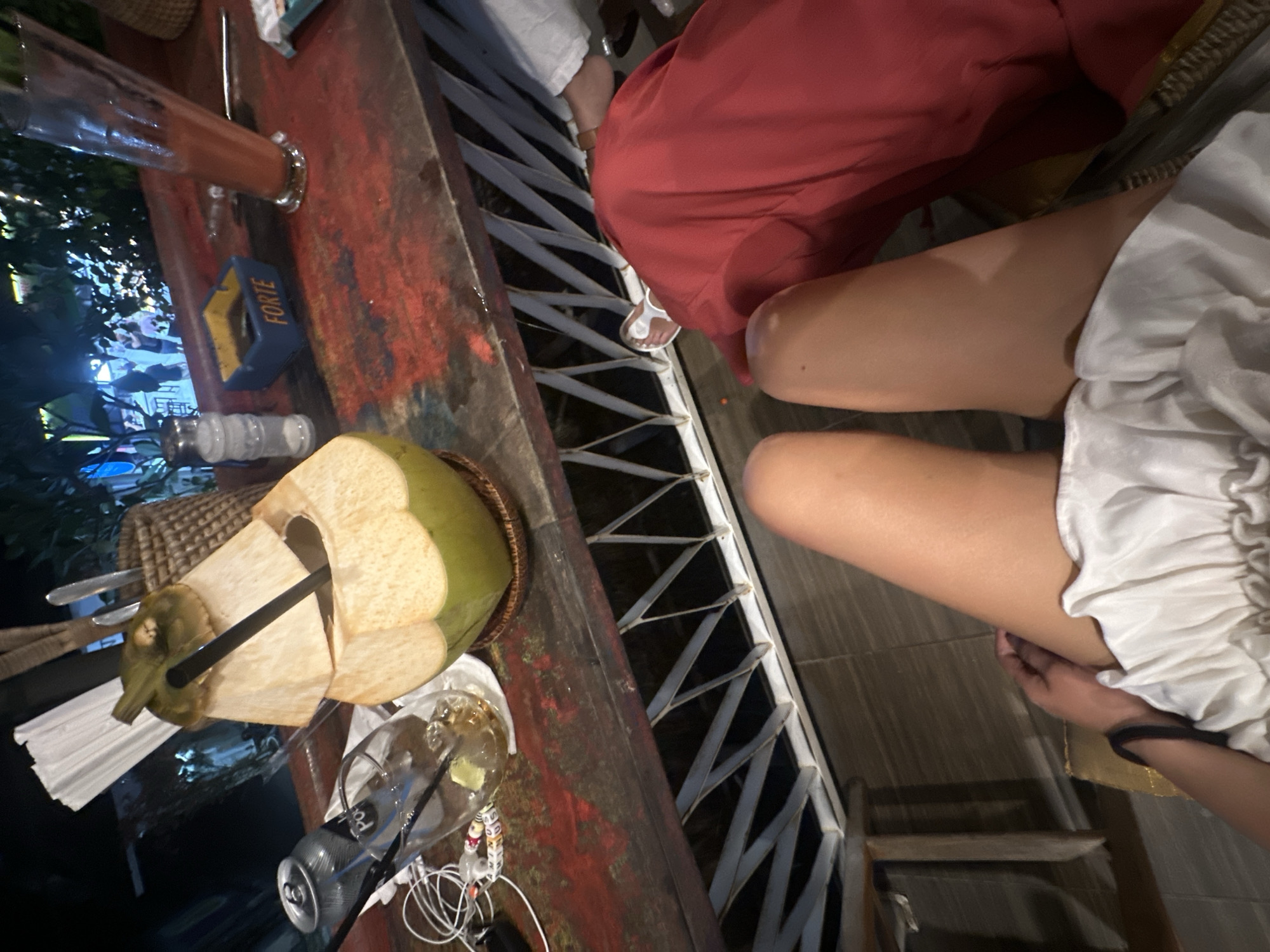 The image depicts a high-angle view of a table setting at a restaurant, possibly in North Kuta, Indonesia, given the presence of local beverages. A partly opened green coconut with two black straws stands prominently on the left side of the red-colored tabletop. Next to it are utensils in a basket and an open can on the table, along with tangled white headphones and a glass with leftover contents. On the same table, a tall glass filled with a red drink is visible. A blue container with the word "FORTE" inscribed on it is also present.
Two individuals are partially visible, seated by the table. One person, dressed in a red garment, is positioned further back, while the other, closer to the foreground, is wearing a white dress. The legs of the second person are visible. A white metal grid-like structure, possibly part of the outdoor seating, stretches across the center of the image. The background suggests an outdoor setting, with glimpses of foliage and lights in the far left. The overall atmosphere indicates a casual dining or social setting.