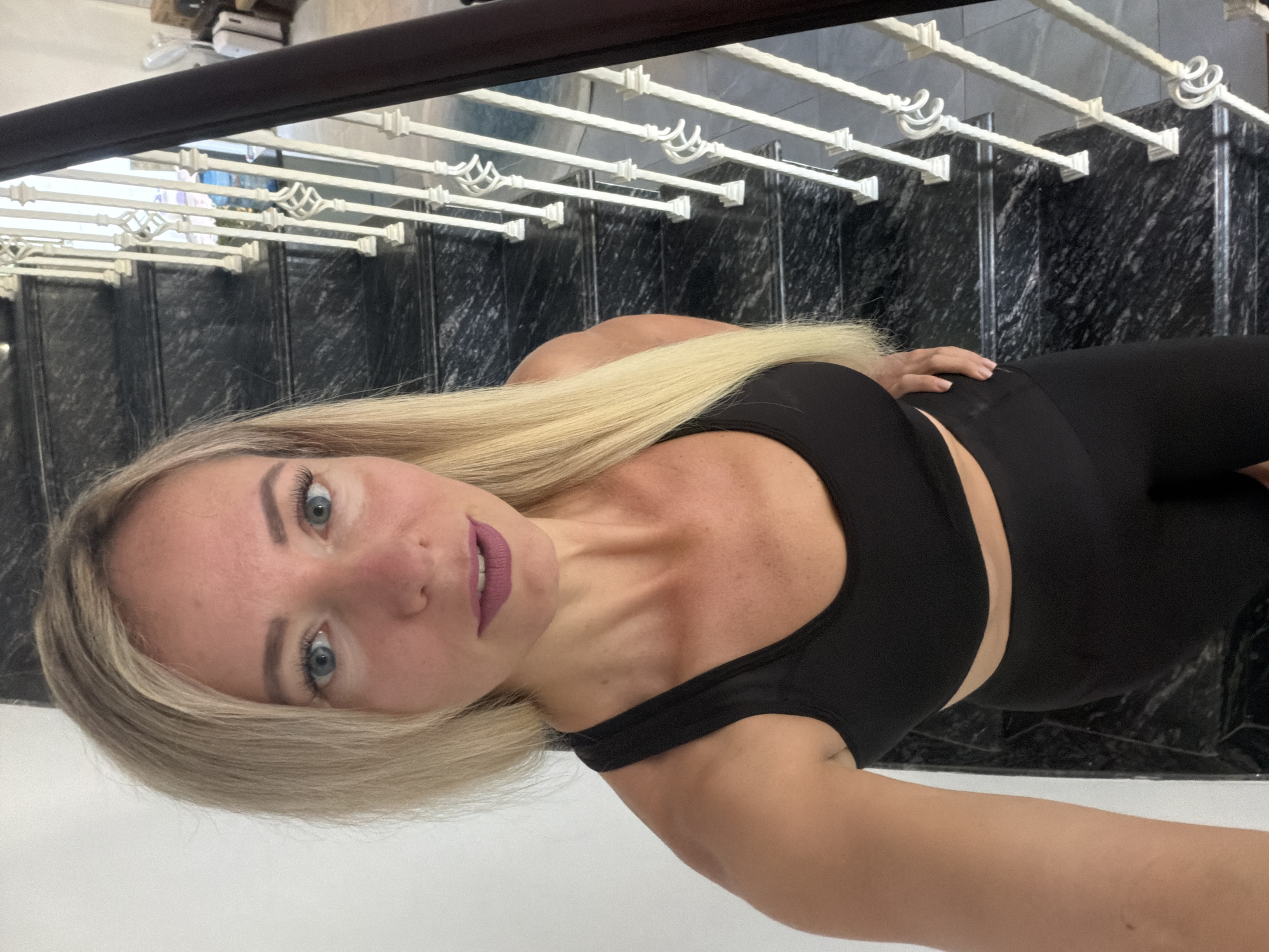 A woman with blonde hair and blue eyes, wearing a black sports bra and black leggings, stands on a marble staircase with white railings. She is looking at the camera and has her left hand on her hip. The location appears to be indoors, possibly a home or a hotel, given the presence of luggage in the background. The scene is brightly lit.