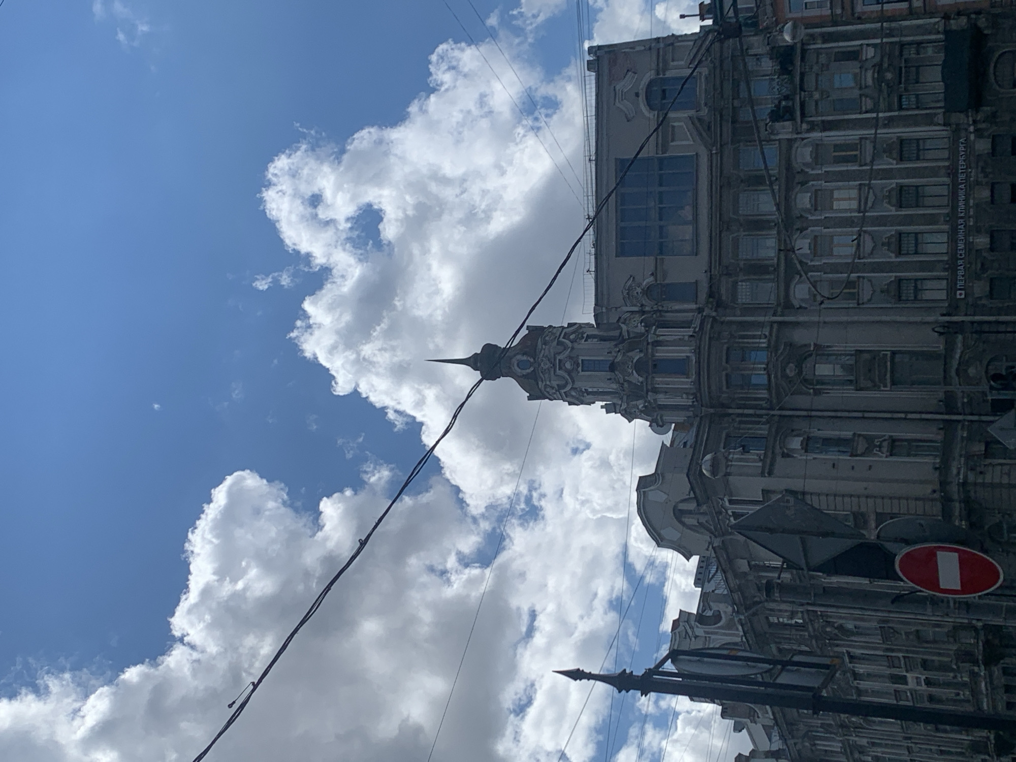 This is a low-angle shot of an ornate, multi-story building in Saint Petersburg, Russia, under a bright, cloudy sky. The building exhibits classical architecture with detailed facades, including pilasters, arched windows, and decorative molding. A prominent tower with a spire rises from the center of the structure. Power lines are strung across the sky, partially obscuring the building and clouds. In the foreground, traffic signs are visible, including a red circular sign with a white horizontal bar, indicating "no entry" or "stop." The building's facade at ground level has a sign in Russian that reads "ПЕРВАЯ СЕМЕЙНАЯ КЛИНИКА ПЕТЕРБУРГА" which translates to "First Family Clinic of Petersburg." The scene depicts an urban streetscape in a historic district.