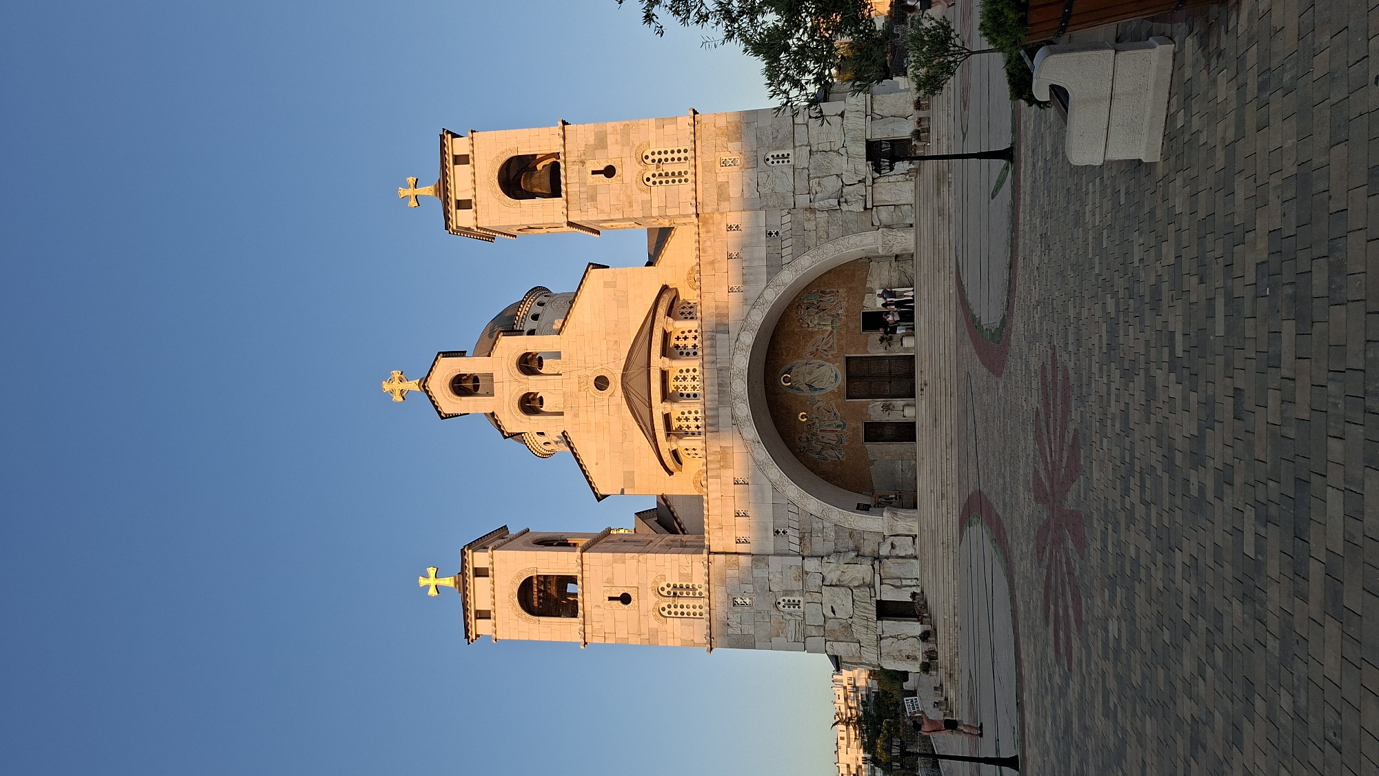 The image shows the exterior of the Cathedral of the Resurrection of Christ in Podgorica, Montenegro. The cathedral is a large, imposing stone building with a central dome and two bell towers. The facade features an arched entrance with mosaics above it. Golden crosses adorn the tops of the bell towers and the dome. The cathedral is bathed in the warm light of the setting sun, casting long shadows across the plaza in front of it. The plaza is paved with cobblestones and features decorative patterns. Several people are visible, including a woman standing on the steps of the cathedral and others in the plaza. A lamppost and a stone bench are also present. The sky is a clear, deep blue.