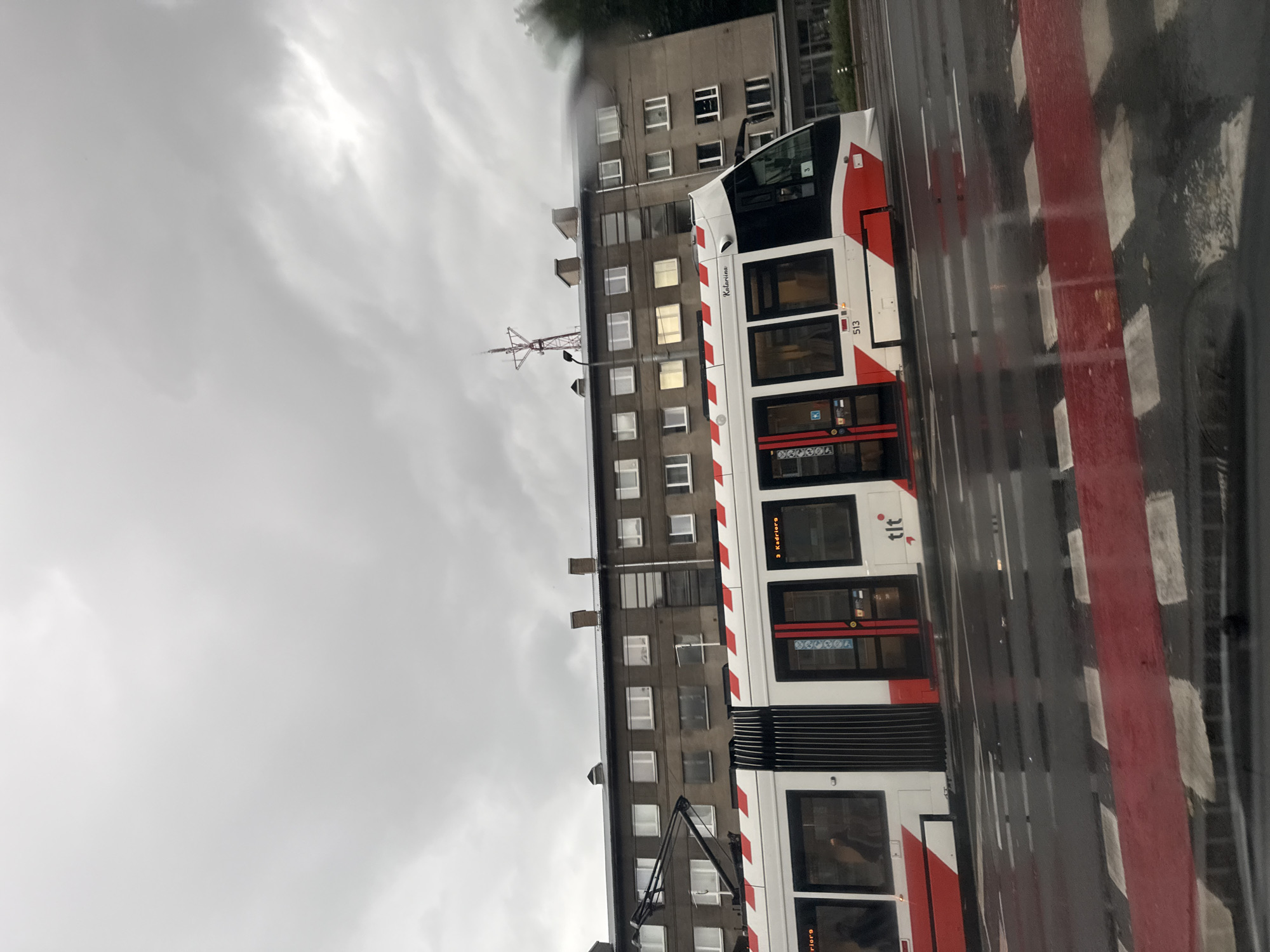 The image shows a modern white and red tram with the number 513 visible on its side, stopped at a crosswalk in Tallinn, Estonia. The tram has the "tlt" logo printed on its side, which is the logo for Tallinna Linnatranspordi (Tallinn City Transport). The tram is adorned with red and white stripes along its body and has "Katarina" written on the top front. The wet road surface indicates it is raining, with reflections visible on the asphalt. The background features a multi-story concrete building with numerous windows, and a cloudy, overcast sky. A tall antenna structure is visible on top of the building to the left of the image. The red and white striped crosswalk is prominent in the foreground.
