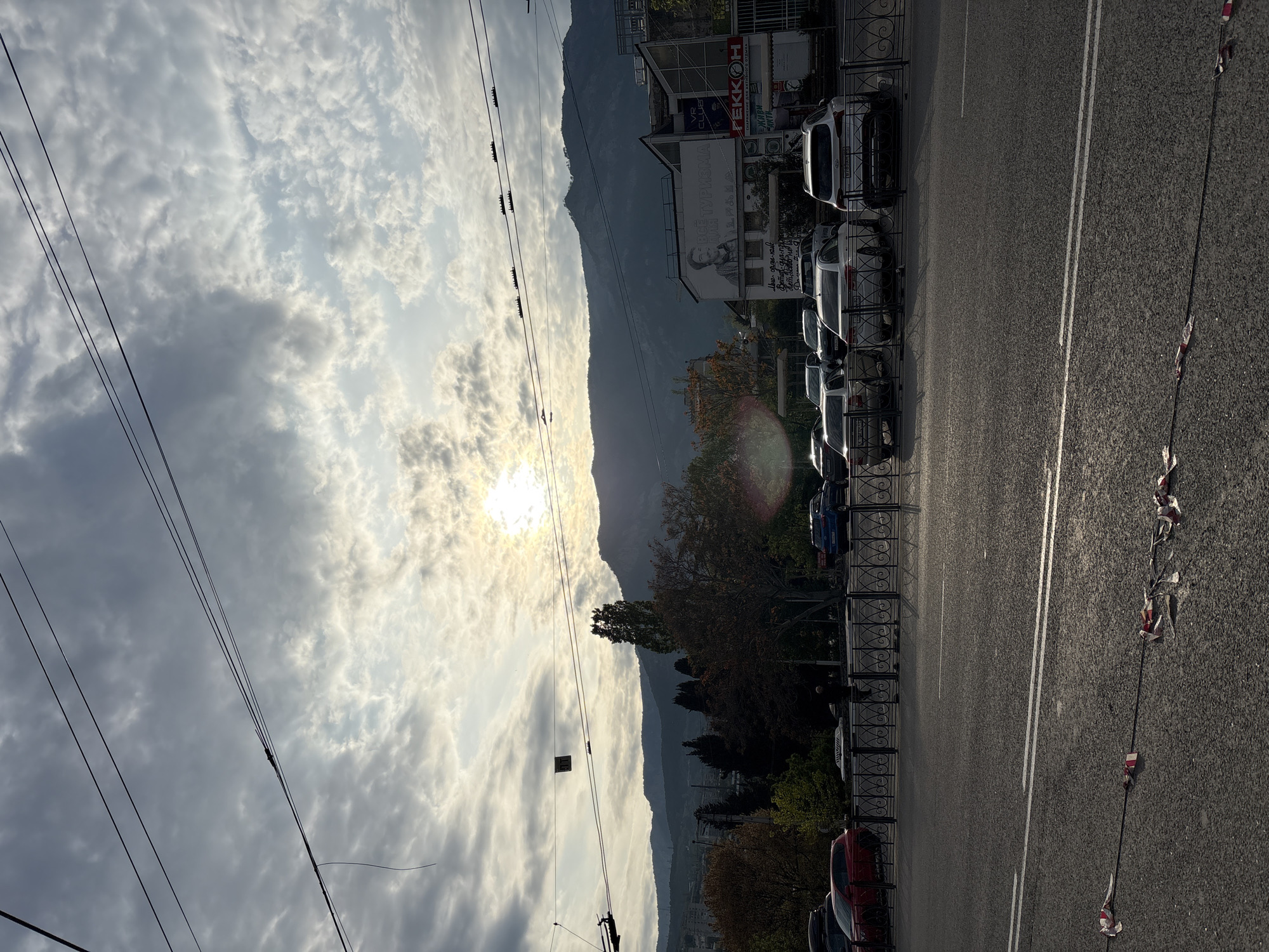 The image shows a street in the city of Ялта, Ukraine, with mountains in the background. The sun is shining brightly through a cloudy sky. A row of parked cars is on the right side of the street, next to a black metal fence. There are signs on the buildings behind the parked cars. A black cable with decorations runs along the road. Power lines are visible in the sky.