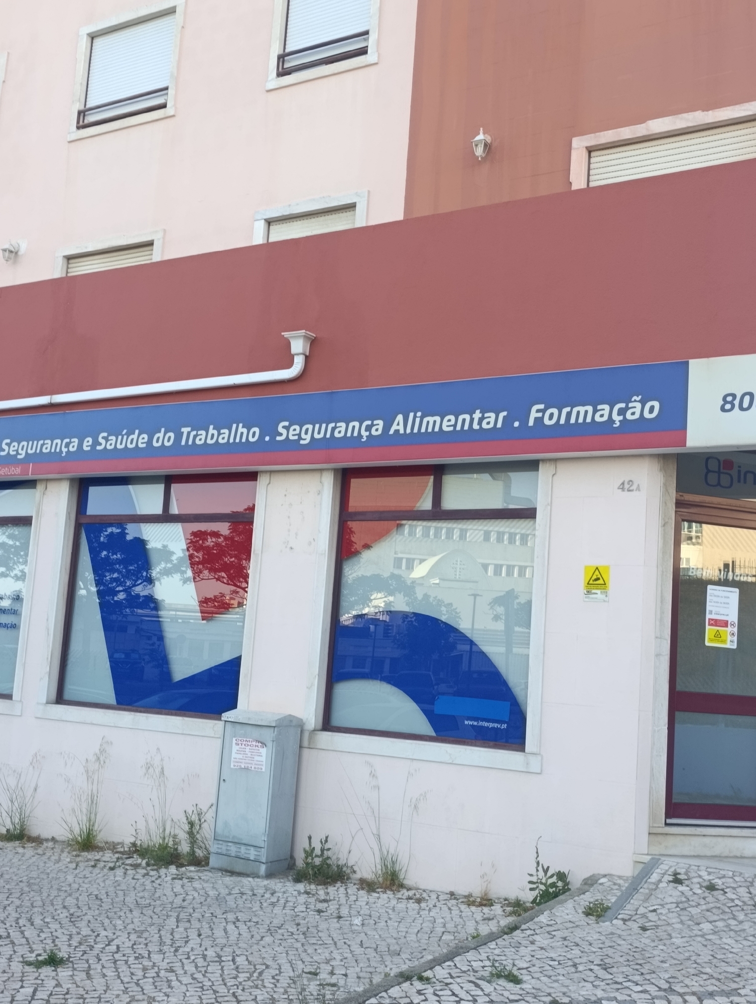 The image shows the exterior of a building with a sign reading "Segurança e Saúde do Trabalho. Segurança Alimentar. Formação." and "Setúbal". The building has large windows, some of which display a blue and red graphic. There is also a white pipe extending from the building. To the right of the entrance, there is a yellow warning sign depicting a falling object. A gray electrical box with a notice on it is situated on the paved ground in front of the building, next to some dry grass. The scene appears to be on a sidewalk or pedestrian area with cobblestone paving.