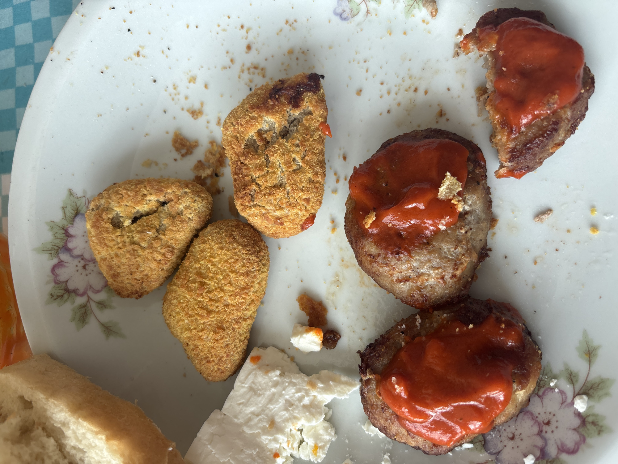 A plate containing food is shown on a table with a blue and white checkered tablecloth. The plate has a floral pattern around the rim. On the plate, there are three breaded, fried food items that appear to have been partially eaten, three meat patties topped with a red sauce, a piece of white cheese, and a piece of bread. The location of Zlatograd, Bulgaria, is not evident in the image.