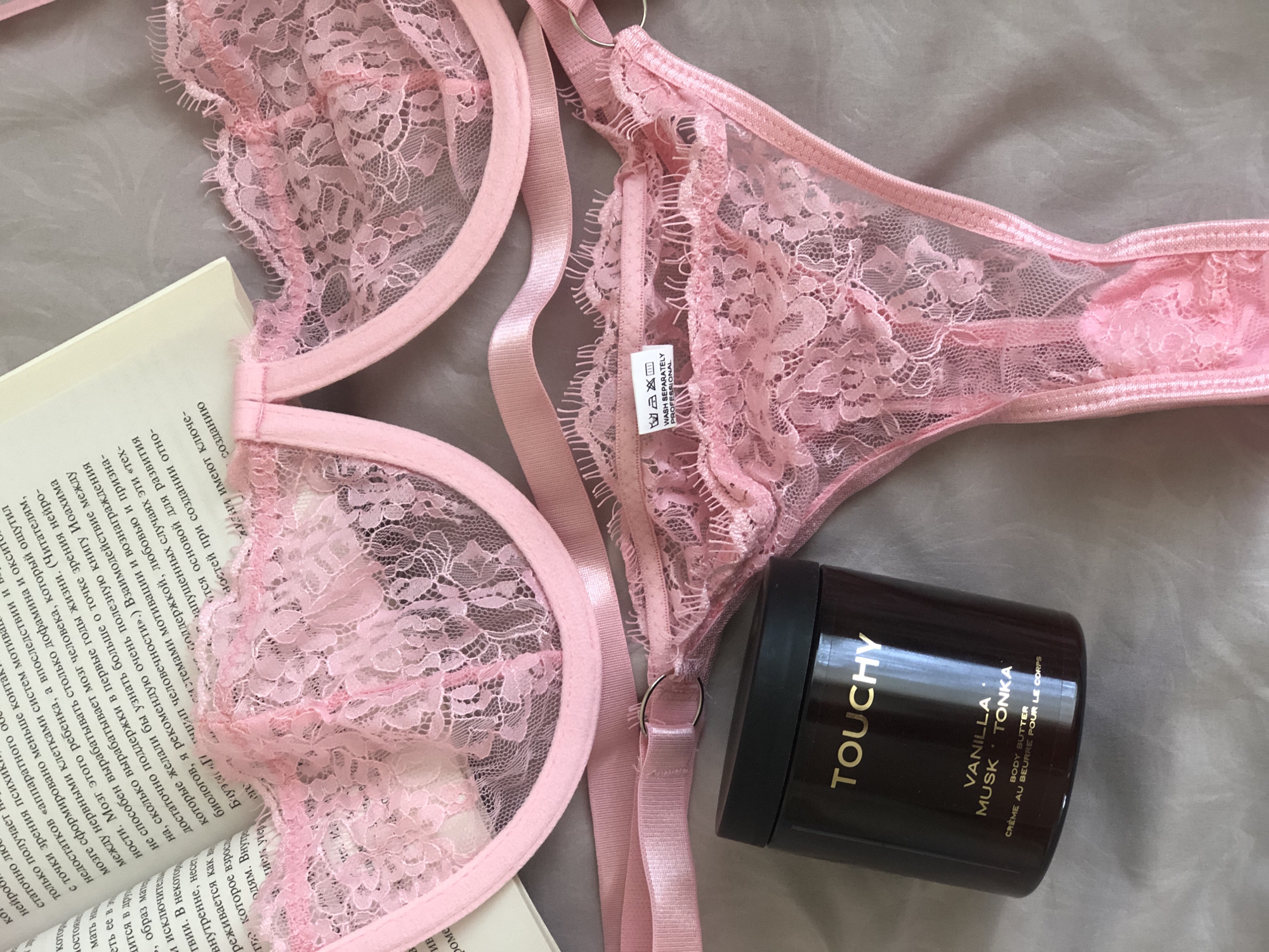 A high-angle close-up shows a pale pink lace bra and matching thong lingerie set lying on a bed or similar surface, accompanied by a book and a jar of body butter. The bra and thong are intricately patterned with floral lace details, and the bra straps are also pale pink. A black jar of "TOUCHY" Vanilla Musk Tonka body butter rests near the lingerie. A partially visible book with pages filled with Russian text is placed on the left. The overall scene suggests a curated or styled aesthetic.