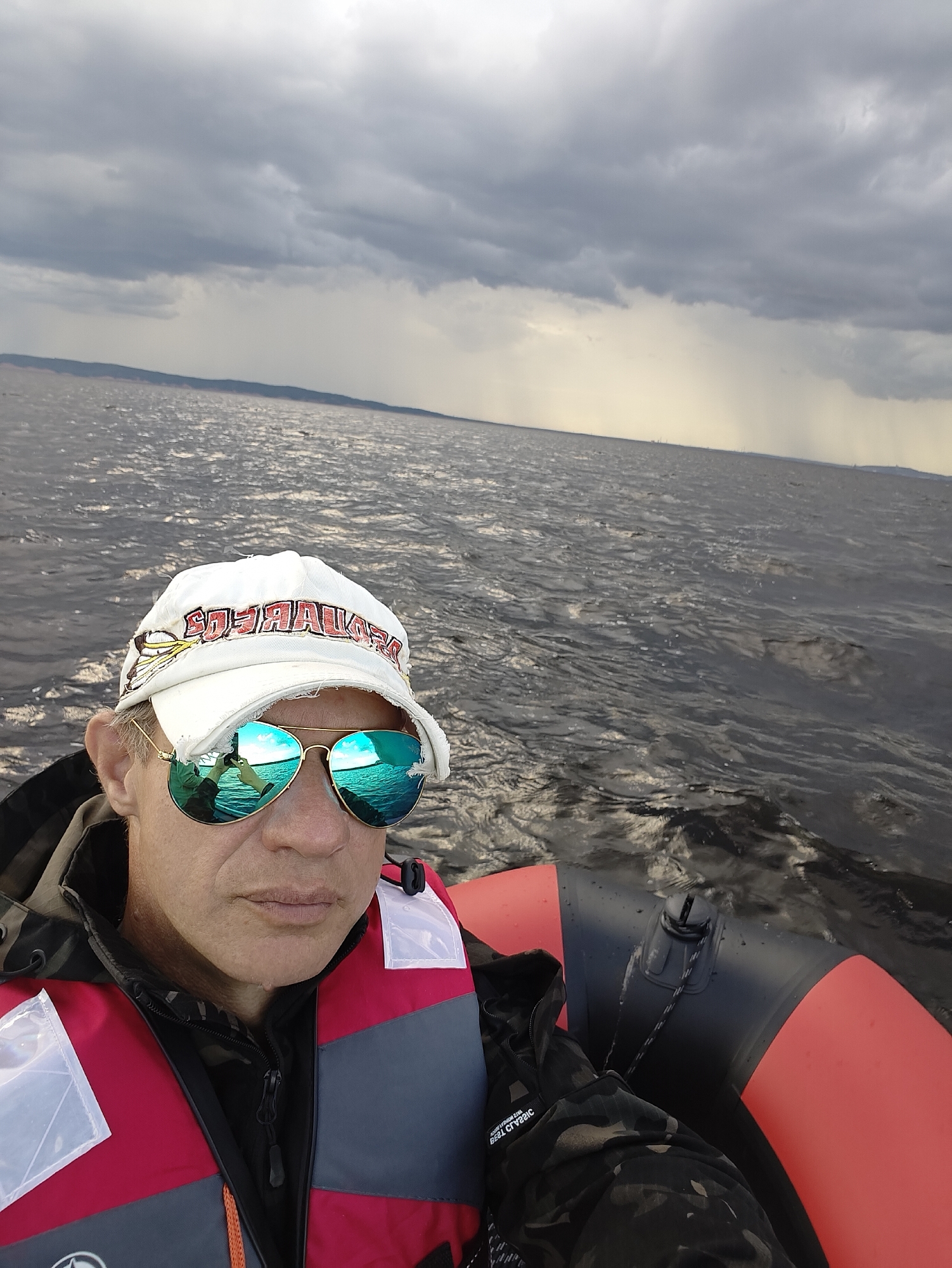 A close-up, front-facing selfie of a middle-aged Caucasian male wearing a white baseball cap and aviator sunglasses, taken from a boat on a body of water. He is wearing a red and gray life vest over a camouflage jacket. The water appears choppy and dark, with a distant, hazy shoreline under a cloudy, overcast sky. A light rain is visible falling in the distance. The man's reflection in his sunglasses shows him holding the camera and another person. The boat appears to be an inflatable dinghy with red and black pontoons. The location is the Kama River in Chaykovsky, Russia.