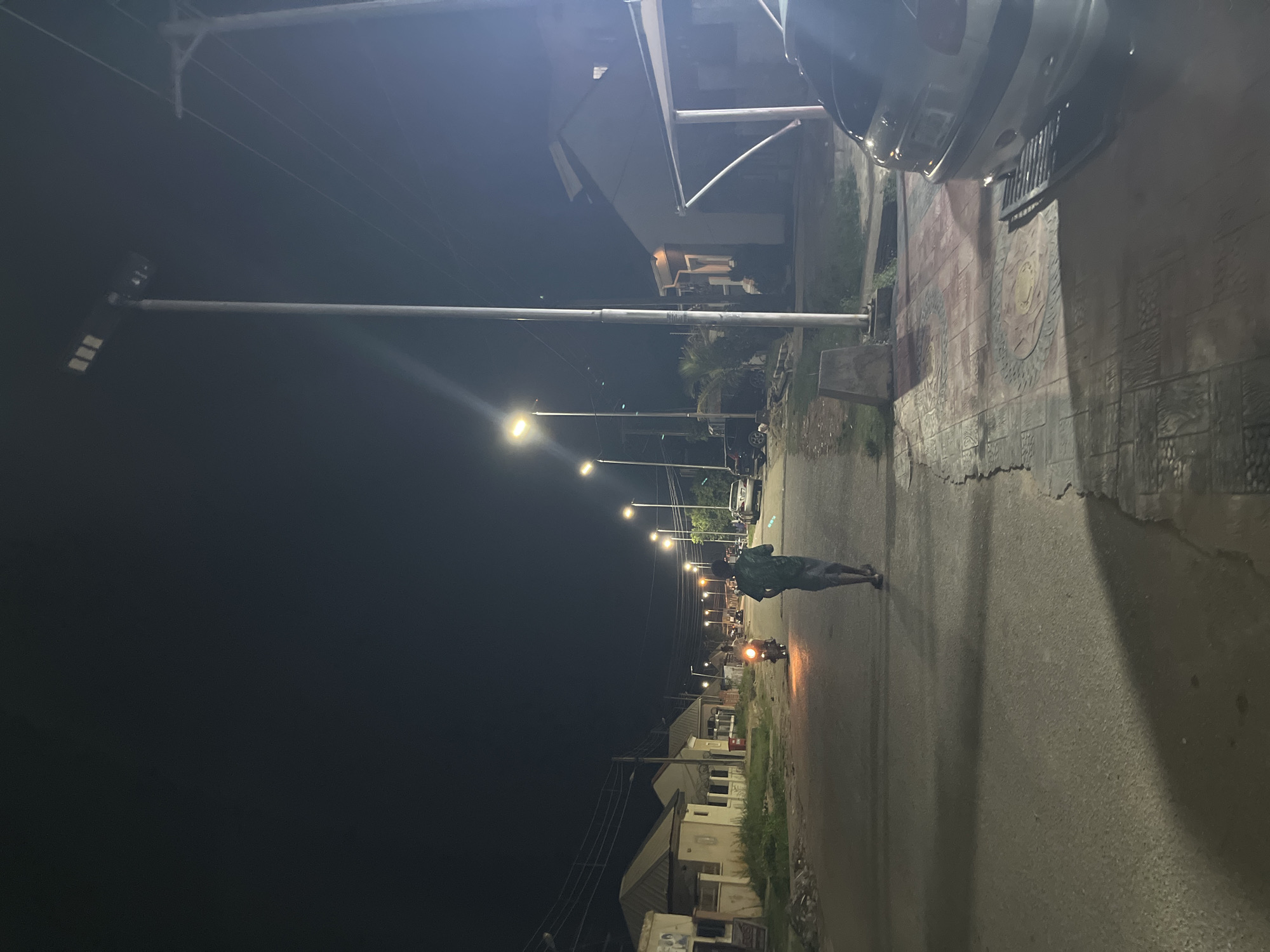 The image depicts a street scene at night in the city of Abuja, Nigeria. Several modern streetlights illuminate the asphalt road, casting long shadows. On the right side of the road, a parked silver car is partially visible. A man is walking away from the camera down the middle of the street. Further down the road, a motorcycle with an illuminated headlight approaches, with a rider visible. On the left side of the street, there are residential buildings with pitched roofs, and some vegetation. Power lines are visible stretching across the dark sky. The overall impression is of a well-lit but quiet urban street at night.