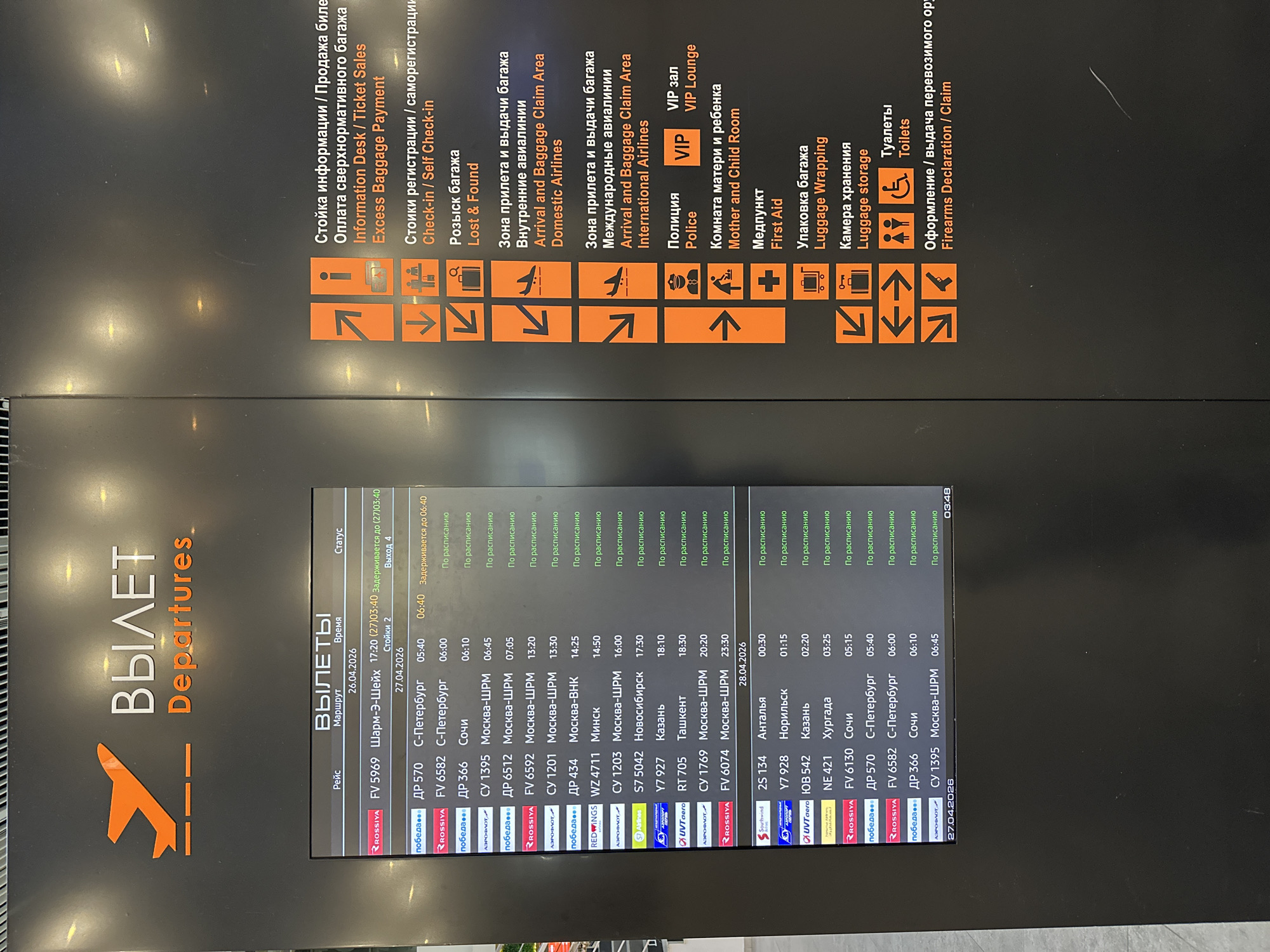 The image depicts a flight departures board in Bolshoye Savino, Russia. The board displays flight information for both domestic and international destinations. Information includes flight numbers, destinations, scheduled times, and status updates such as "Delayed" or "On Schedule." Logos of various airlines are visible next to the flight information. The upper portion of the image shows symbols for airport amenities, including information desks, check-in, lost and found, arrival/baggage claim, police, first aid, luggage services, toilets, and firearms declaration. The departures board includes flights scheduled for both April 26, 2026, and April 27, 2026.
