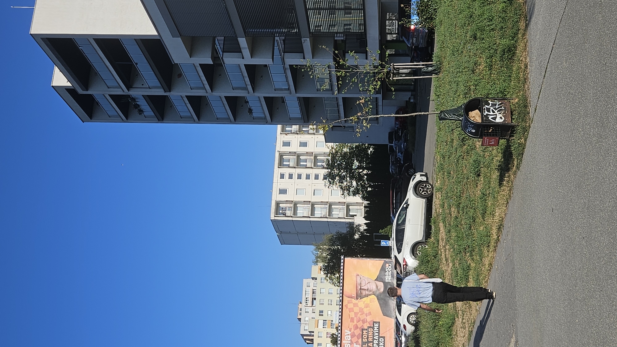 The image shows a street scene in the city of Prešov, Slovakia, on a clear, sunny day. In the foreground, a man wearing a blue t-shirt and dark pants walks away from the camera on a paved walkway. He carries a white tote bag over his left shoulder. To his right, a patch of green grass slopes upwards, dotted with dandelions and other weeds. A black trash can, adorned with white graffiti, sits on the grassy area.
Behind the grassy slope, several cars are parked. A white SUV is prominently visible in the middle ground, with other vehicles, including a white sedan and darker cars, in the background.
Further back, an outdoor advertisement billboard features a portrait of a man wearing an orange hard hat, with text in Slovak. Behind the billboard and the cars, multiple apartment buildings are visible. The building closest to the viewer on the right is a modern, multi-story apartment complex with balconies and glass railings. To the left of this building, a taller, older apartment building with rows of windows and balconies stands. In the distance, another apartment building is visible. The sky is a clear, vibrant blue with no clouds. The overall scene depicts a typical urban environment with residential buildings and public spaces.