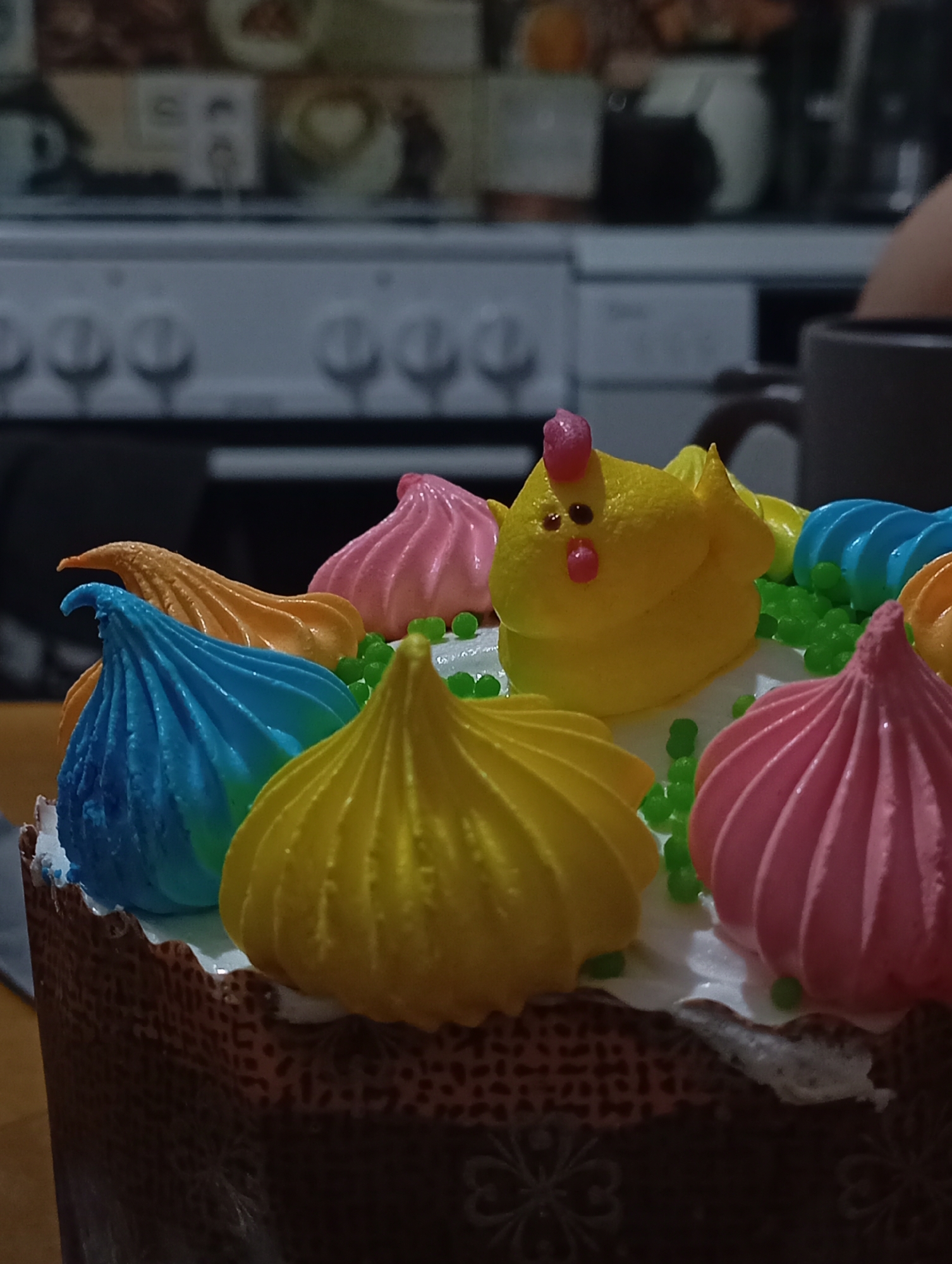 A close-up shot of a decorated Easter cake, likely a Kulich, sits on a wooden surface. The cake is adorned with white frosting and colorful meringue decorations in the shape of chickens and teardrops, with green sprinkles between the meringues. Colors of the meringues include yellow, pink, blue, and orange.
In the blurred background, there is a kitchen stove and cupboard, suggesting that the picture was taken in a domestic setting. There is a cup next to the cake.
The information provided does not contain information about the location in the city of Krasnodar, Russia.