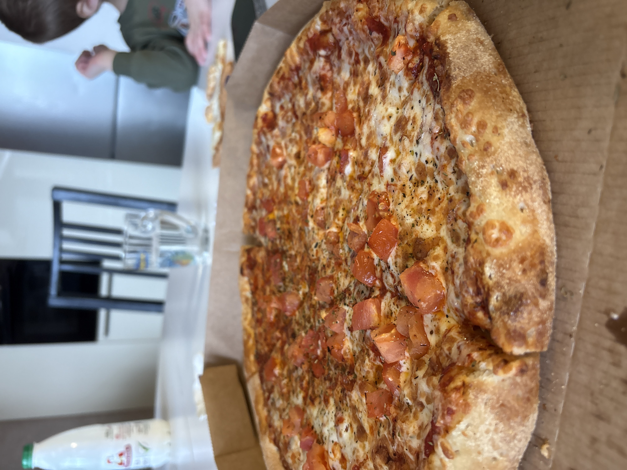 The image captures a close-up of a large, round pizza served in a cardboard delivery box, placed on a table in an indoor setting, likely a kitchen or dining area. The pizza dominates the right side of the frame, presented from a slightly elevated, side-on perspective. It features a thick, golden-brown crust, generously topped with melted cheese, numerous diced red tomatoes, and a sprinkling of dark herbs, possibly oregano or basil. The pizza appears to be cut into slices.
To the left of the pizza, the top of a child's head and a portion of their upper body, wearing a green long-sleeved shirt, are visible in the upper left corner. The child is out of focus, suggesting they are a secondary subject or in the background, possibly reaching towards the table or pizza.
On the table in the lower-left corner, a white plastic bottle with a green cap is visible, alongside a clear glass. The bottle has a red design and some indistinct Cyrillic text, including what appears to be the brand "Агуша" (Agusha). The background reveals blurred elements of a domestic interior, possibly white cabinets or walls, and what might be a dark appliance or furniture. The lighting suggests it is daytime. The overall scene conveys a casual mealtime atmosphere.