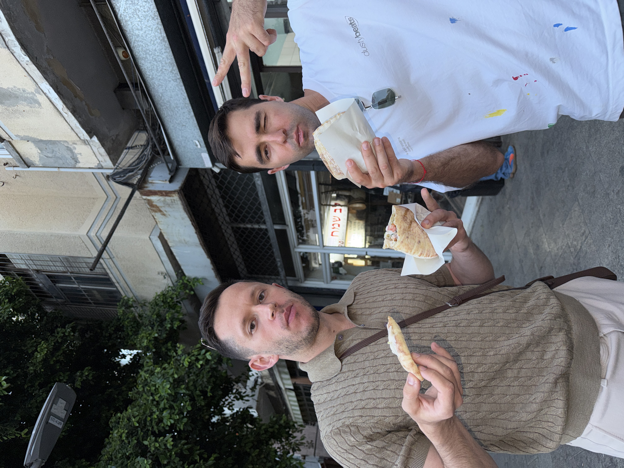 Two men are standing on a city street. One man is wearing a white t-shirt and the other is wearing a brown sweater. Each man is holding food in his hands. They are standing near a building with a sign in Hebrew. The location is likely in Tel-Aviv, Israel.
