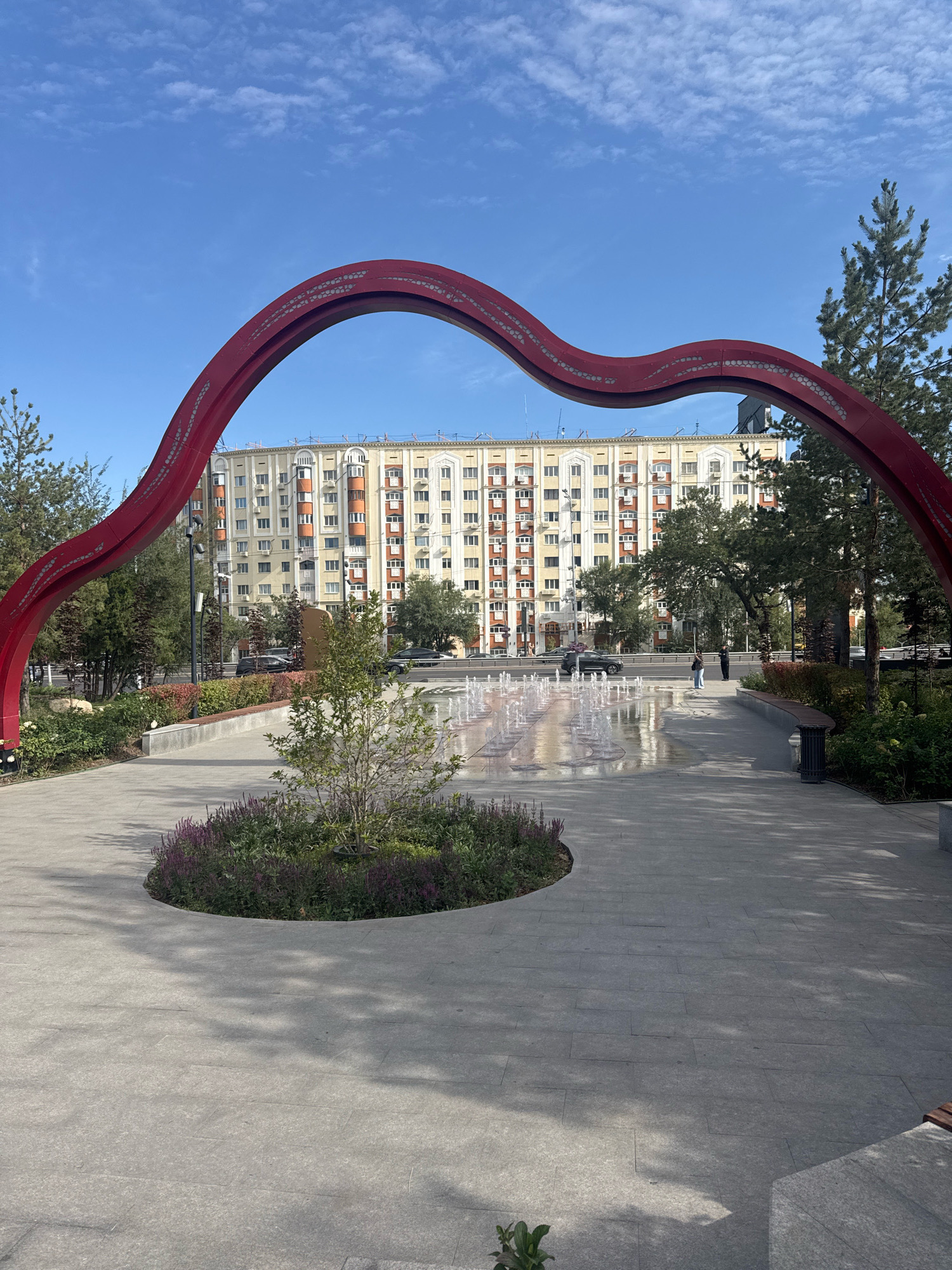 The image captures a public plaza in Almaty, Kazakhstan, featuring a distinctive red, wavy archway as a central art installation. The archway, adorned with a pattern of small circular openings, frames a view of a large, multi-story apartment building with a light yellow facade and numerous windows. In front of the building, a modern plaza unfolds. A shallow water fountain with numerous jets creates a shimmering effect on the paved surface. Lush green trees and smaller decorative plants, including a bed of purple flowers surrounding a small tree, add to the urban greenery. A paved walkway leads towards the fountain area, and a dark-colored trash receptacle is visible to the right of the pathway. Two individuals are seen walking in the distance, near the fountain. The sky above is a clear blue with scattered white clouds. The scene appears tranquil and well-maintained, typical of a public recreational space.