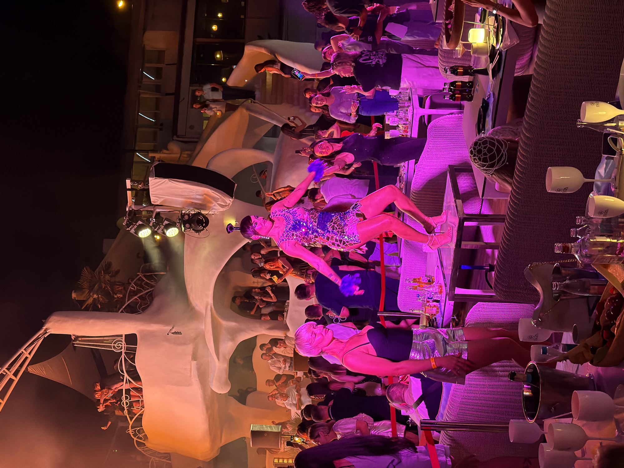 This image depicts a lively nightclub scene at night, illuminated by vibrant pink and orange lighting. In the foreground, a woman with short blonde hair, wearing a black sequined top and silver shorts, stands among a crowd of people. To her right, a performer in a sparkly, patterned dress is dancing on a raised platform, holding a blue feathered object. In the background, various individuals are seen socializing, some holding phones and taking pictures. The architecture of the club appears to be organic and sculptural, with white, flowing structures and illuminated panels. Tables are set with drinks and food, and lounge seating is visible.
The specific location of this nightclub is the city of Odesa, Ukraine.