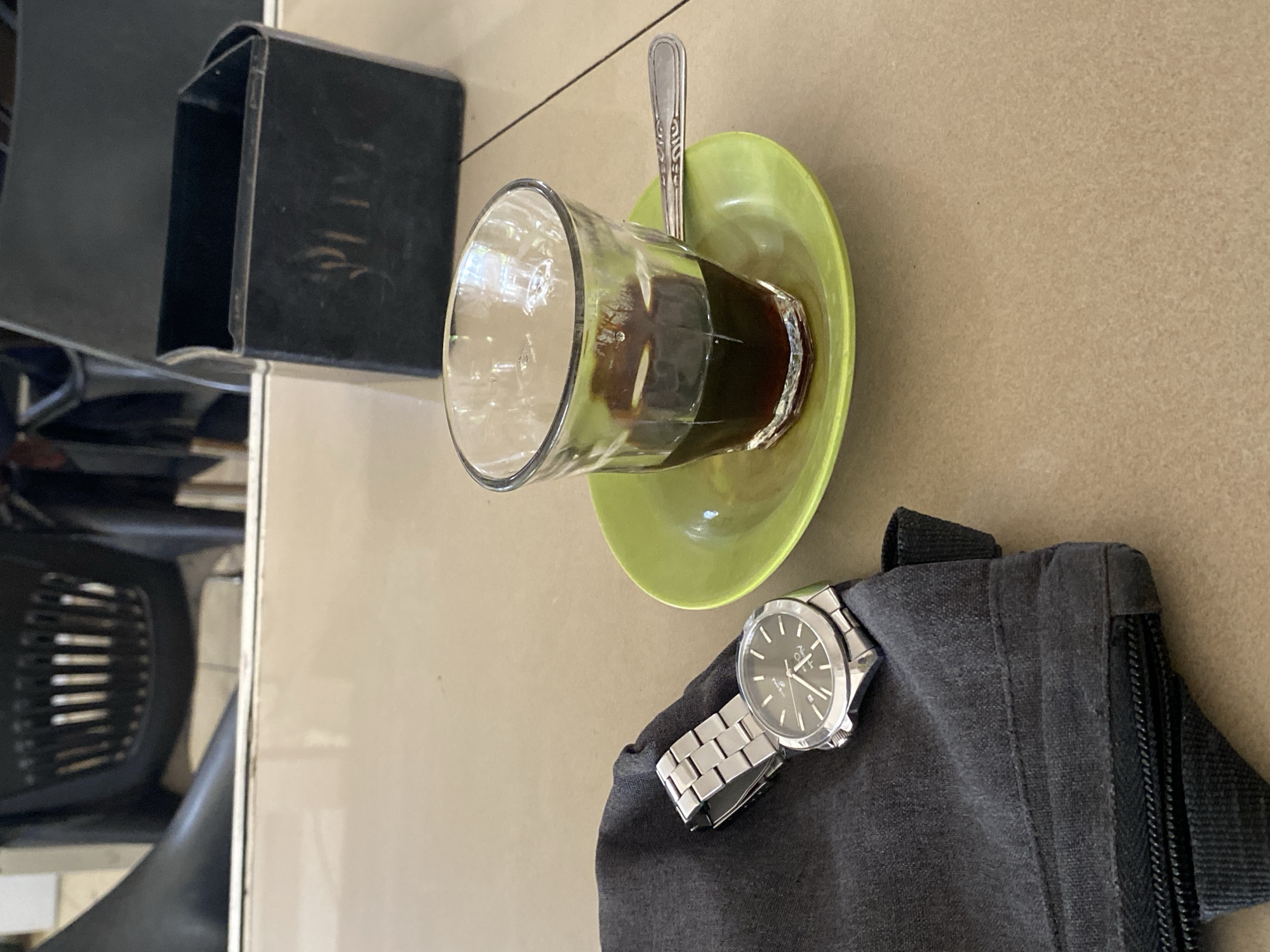 An overhead, medium shot captures a table setting at what appears to be a cafe in Lhokseumawe, Indonesia. In the foreground, a dark grey fabric pouch with a zipper closure rests on the table. A silver wristwatch with a black face is laid on top of the pouch, its metal band partially visible. To the right of the pouch, a small, lime-green saucer holds a clear glass filled with dark coffee. A silver spoon is placed across the saucer and into the glass. In the background, a black container with the word "SLIM" faintly visible on its side sits on the table. Behind that, out-of-focus black chairs are visible. The table's surface is a light beige color.