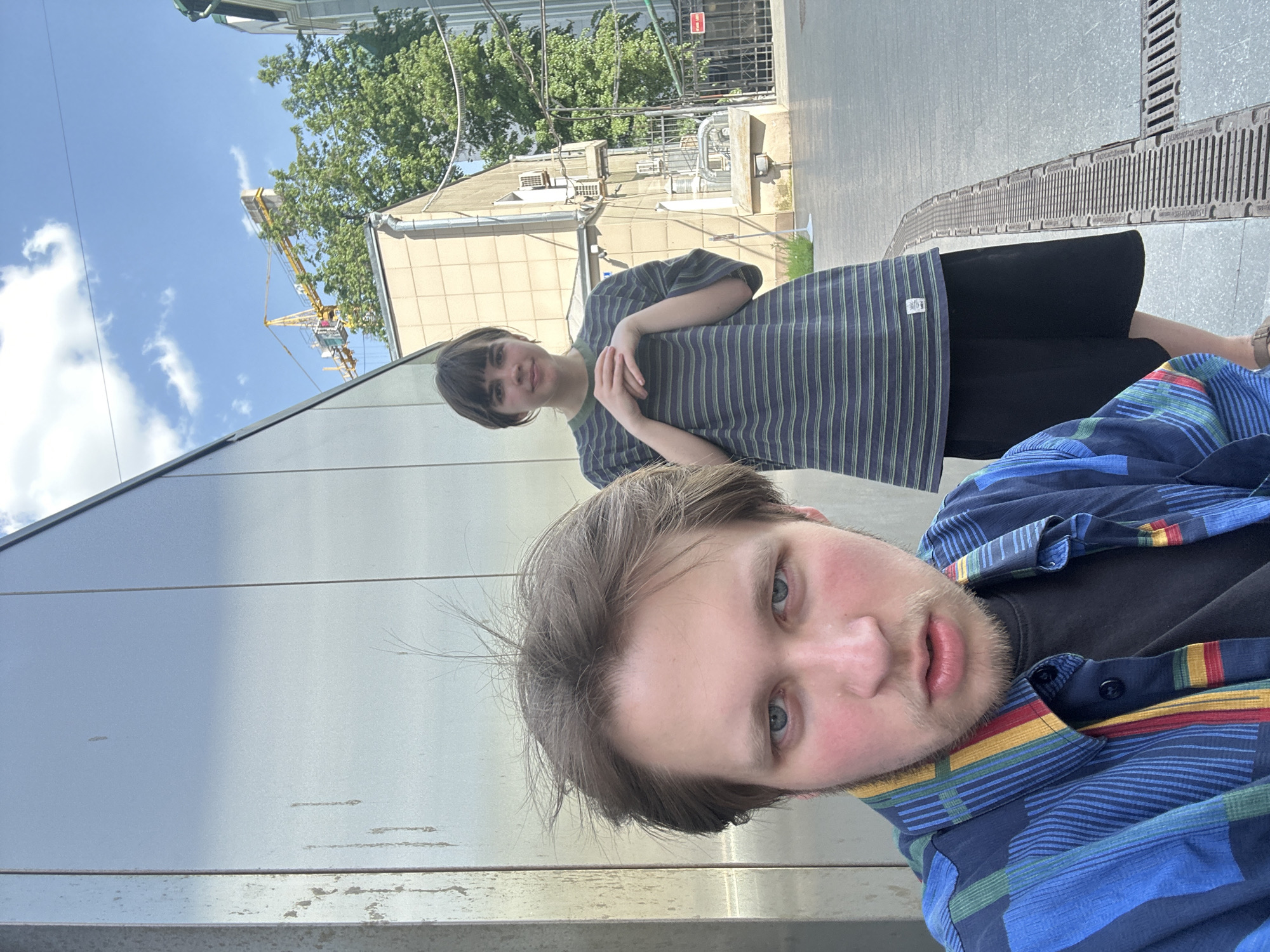 Two individuals are pictured outdoors in an urban environment. The foreground features a person with short brown hair and blue eyes, wearing a black t-shirt and a blue, yellow, and red plaid shirt. The person's face is close to the camera, creating a selfie-style perspective.
Behind them stands a second person with dark hair and a striped shirt. Both individuals appear to be on a sidewalk or pedestrian area, with a metal grate visible on the right side of the frame.
In the background, there is a building with a crane visible in the distance. The sky is blue with some clouds.
