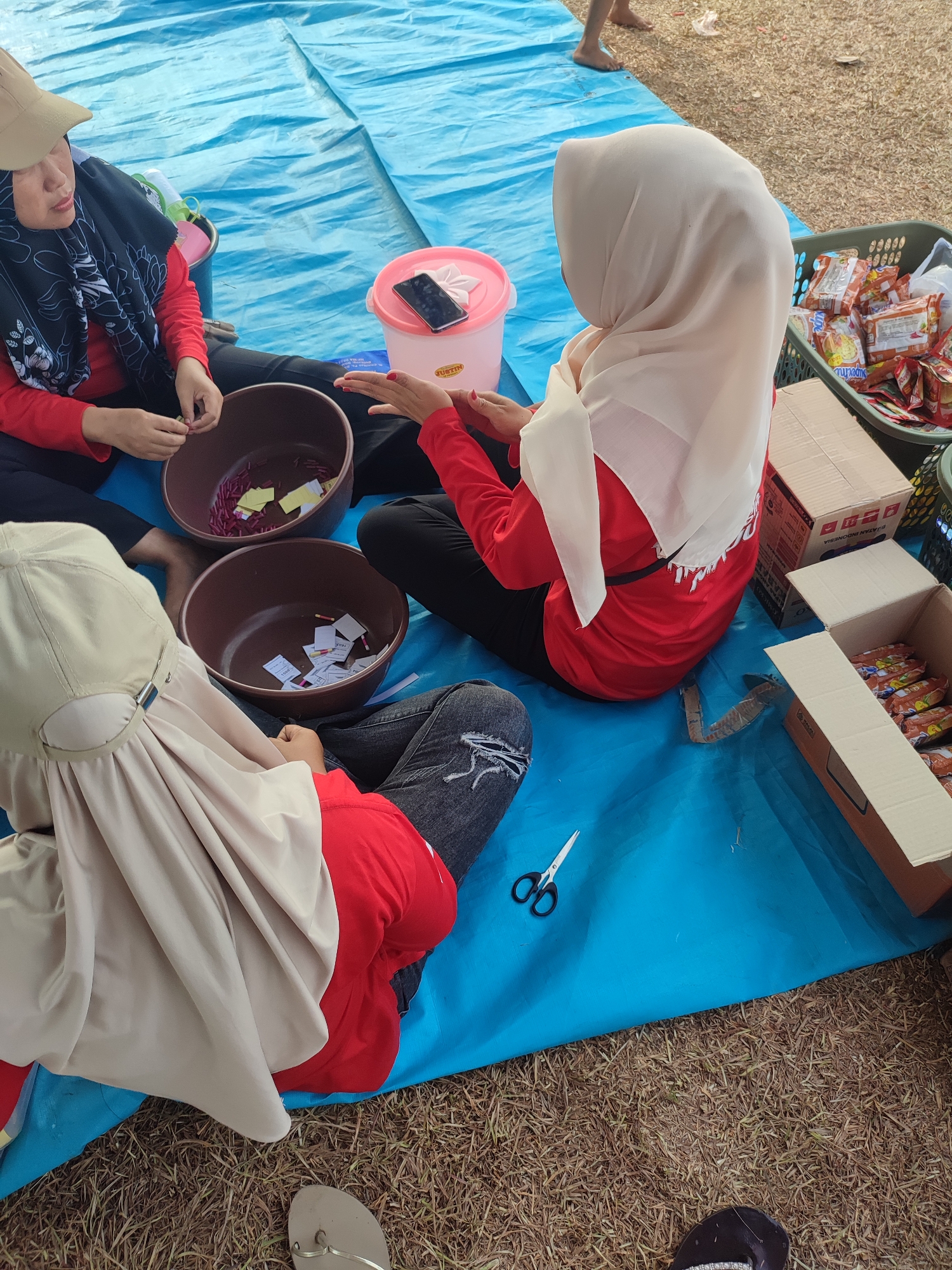 The image depicts an outdoor scene on a bright day, likely in Nusa Jaya, Indonesia. Three individuals are present, seated on a large blue tarp spread over dry grass. Two of the individuals are women wearing red shirts and headscarves. The woman on the left wears a tan cap over her headscarf, and the woman on the right wears a light beige headscarf. The third individual, positioned in the foreground, is partially obscured, but appears to be wearing a red shirt and a tan head covering.
In front of the seated individuals are two dark brown bowls containing small, rolled pieces of paper, likely for a lottery or game. A pink plastic container with a phone resting on top is situated near the woman on the left. To the right of the group, several cardboard boxes and a green plastic basket filled with packaged food items, possibly instant noodles, are visible. A pair of scissors lies open on the blue tarp near the foreground. The overall impression is of a gathering or event where participants are engaged in preparing or drawing from the paper rolls.