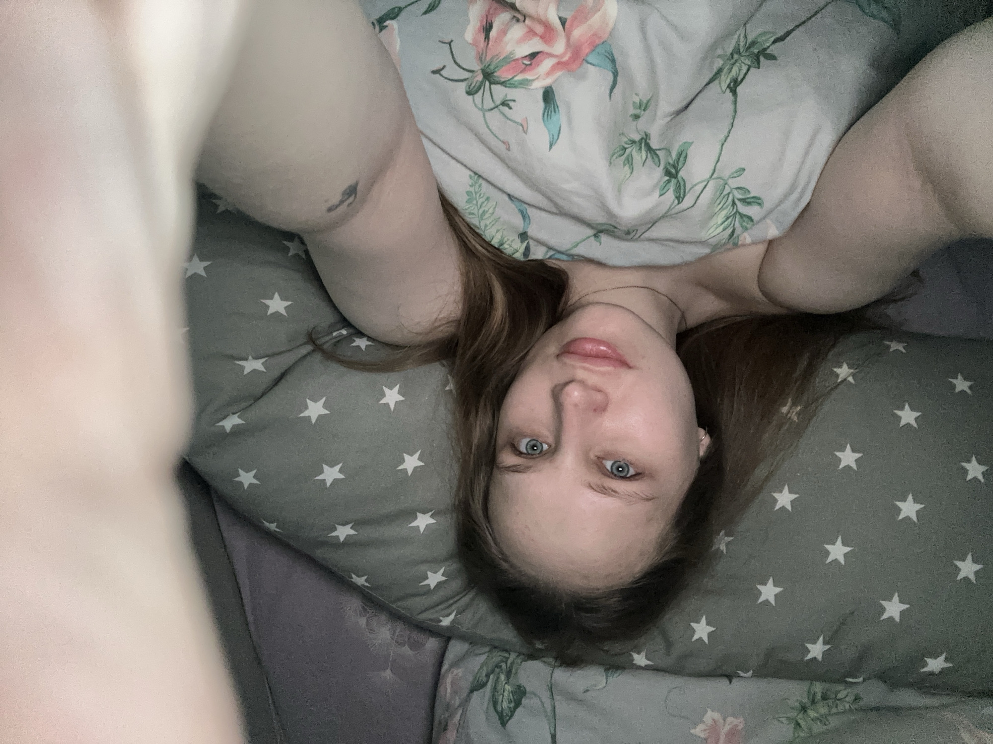A woman with light skin and blue eyes lies on a pillow with white stars on a gray background. Part of a floral patterned blanket covers her upper body. The photo is taken from an overhead perspective. There is no evidence to indicate the location is Chaikovsky, Russia.