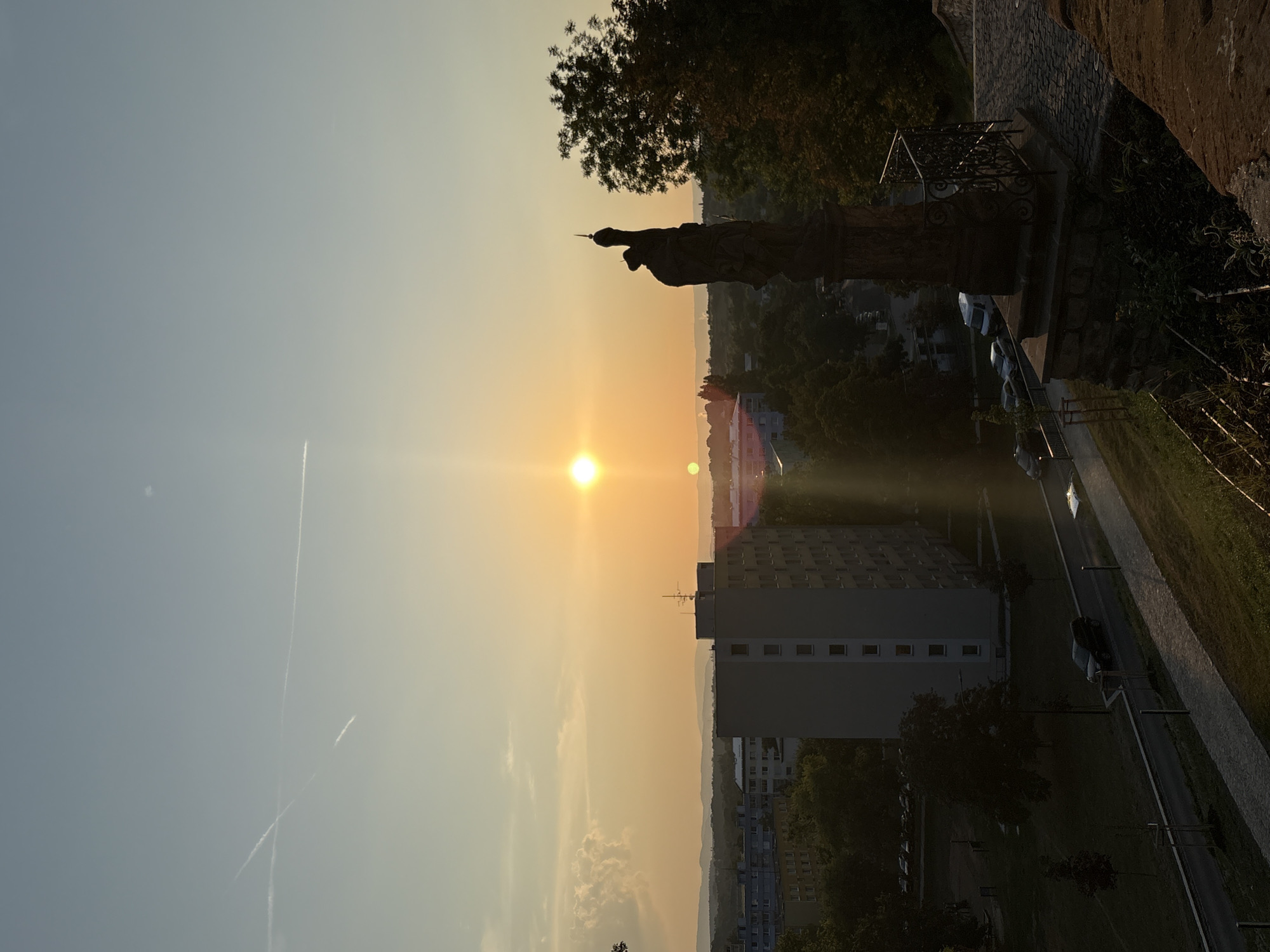 This is an image taken at sunset, looking out over a city. The sun is low in the sky, casting a warm orange glow. In the foreground, there is a stone statue of a person. Behind the statue, there are trees and buildings. A tall, light-colored apartment building is prominent in the mid-ground. There are several cars parked on the street below the apartment building. The sky is clear with a few wispy clouds. There are also contrails from airplanes visible in the sky. The image appears to have been taken from an elevated position, possibly a hill or a tall building overlooking the city.