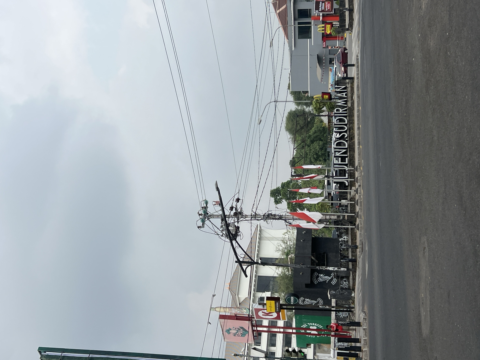 The image captures a street scene in Yogyakarta, Indonesia, featuring a large monument with the text "JL. JEND. SUDIRMAN" in prominent white lettering against a dark background. Several Indonesian flags are displayed on flagpoles in front of the monument, adding a patriotic element to the scene. To the right of the monument, a McDonald's restaurant is visible, indicated by its iconic golden arches logo and red signage.
The surrounding environment shows a mixture of buildings and infrastructure. On the left, a tall green structure with text and what appears to be a bottle graphic stands out, possibly a billboard or part of a shop. A traffic light with a green signal is also visible. Further back, white buildings with distinctive rooftops suggest institutional or commercial establishments. Electrical poles with a complex network of wires are a common sight, indicating the urban setting. The road in the foreground is asphalt, and there are indications of a sidewalk and street furniture like bollards.
A few vehicles are present, including a red car driving near the McDonald's entrance. A person in a red shirt and beige pants is standing near the green structure on the left, appearing to be an observer or patron. The sky is overcast, with a diffused light suggesting a cloudy day. The overall impression is a busy urban intersection in Yogyakarta, marked by prominent landmarks and commercial activity.
