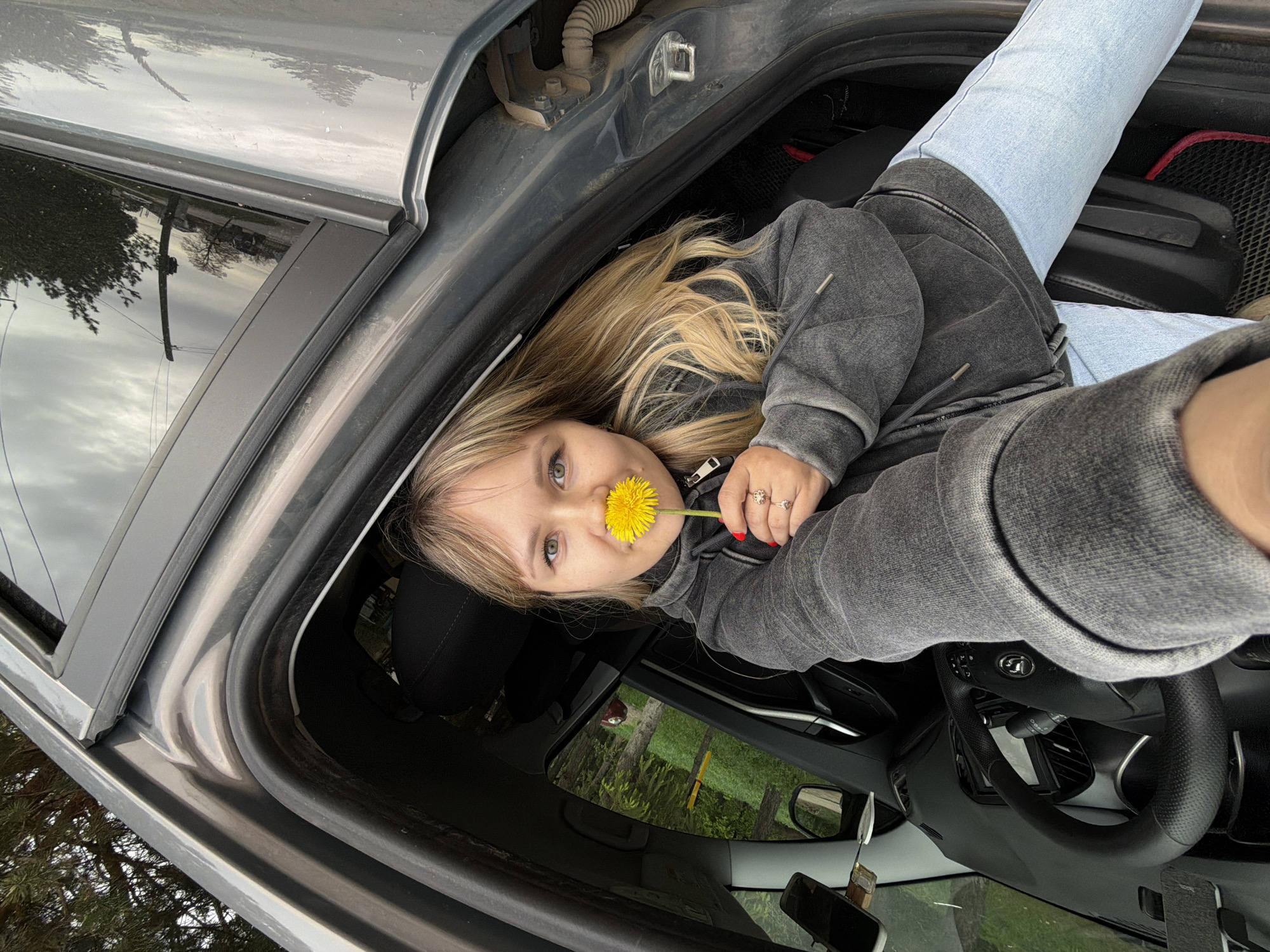 A woman with blonde hair is inside a gray car, holding a yellow dandelion flower to her nose. She is wearing a gray jacket and light blue jeans. The car appears to be parked outdoors, with glimpses of trees and greenery visible through the windows. Based on the information provided, the location is Chaikovsky, Russia. The car's interior is visible, including the steering wheel, dashboard, and seats. The woman is positioned in a way that suggests she might be in the driver's seat or leaning into the car from outside.