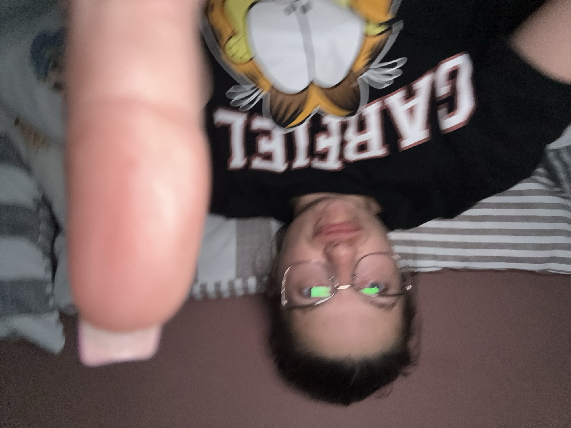 A selfie shows a person lying on a bed with striped sheets. The person is wearing a black Garfield T-shirt and glasses. A blurred finger is in the foreground. The location cannot be determined from the image.