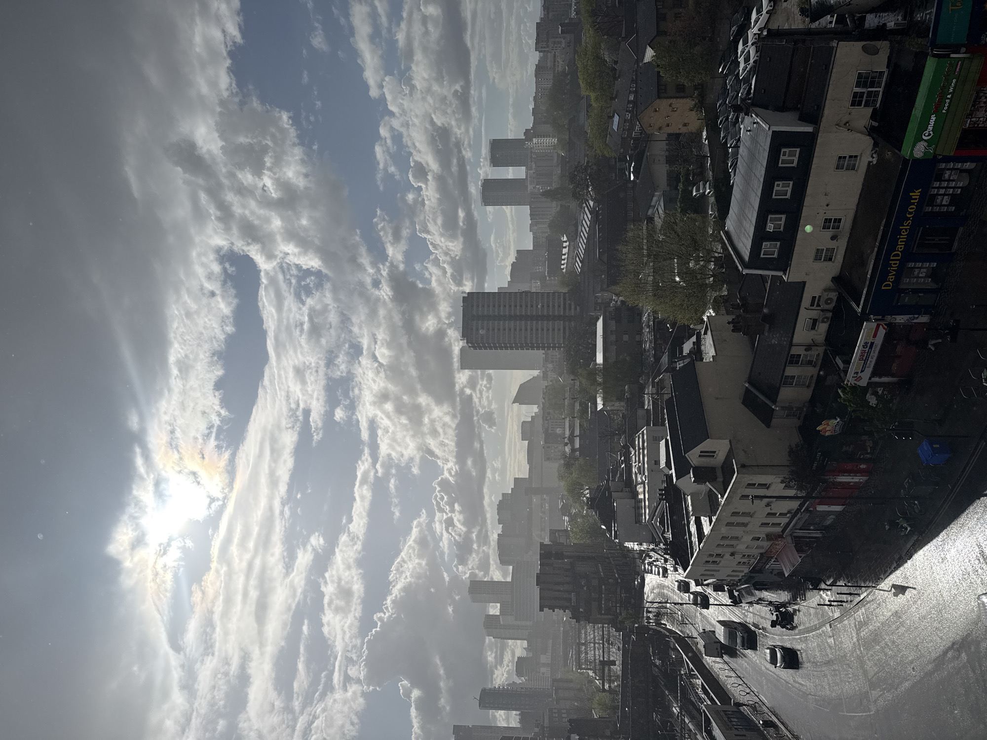 The image captures an elevated view of an urban area, characterized by a mix of residential and commercial buildings. The weather appears to be overcast, with the sun visible through breaks in the clouds. A wide street lined with buildings is visible in the foreground with several cars driving on it. A sign for "DavidDaniels.co.uk" is visible on one of the buildings. The city skyline is punctuated by several high-rise buildings, suggesting this is most likely a central area of London, United Kingdom.