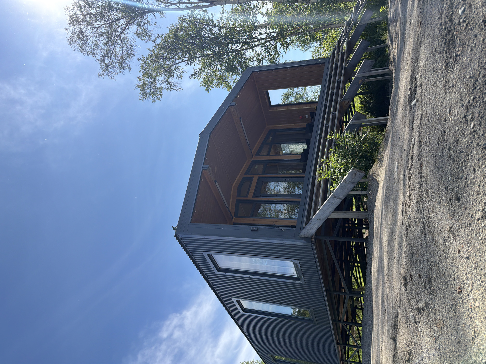 This is a low-angle, exterior, eye-level, full shot of a modern, dark gray corrugated metal house on stilts. The house has several tall, narrow windows and a covered porch with glass doors. The structure is surrounded by trees and sits on a gravelly incline. The sky is clear blue with some wispy clouds and bright sunlight. The location is identified as a city in Russia.