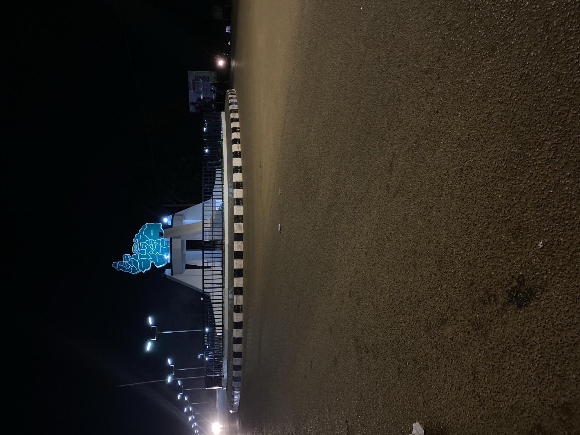 This night-time image captures a quiet street scene, featuring a prominent, illuminated monument. The monument, situated on the left side of the paved asphalt road, is shaped like a geographical map and glows with a distinctive turquoise-blue light. It appears to depict a region or state, intricately divided into smaller administrative areas, with one circular segment clearly marked "CITY."
Alongside the road, a black and white striped curb or barrier separates the roadway from the monument structure. A series of modern streetlights equipped with bright LED lamps line the street, casting an even illumination across the scene. The sky is completely dark, indicating a late hour, and there are no visible people or vehicles, contributing to a serene and deserted atmosphere.
The location context is Bukuru, Nigeria, suggesting the map monument likely represents Plateau State or a specific local government area within it. Beyond the immediate foreground, some indistinct structures and further streetlights recede into the darkness. The weather appears clear, with no signs of rain or fog.