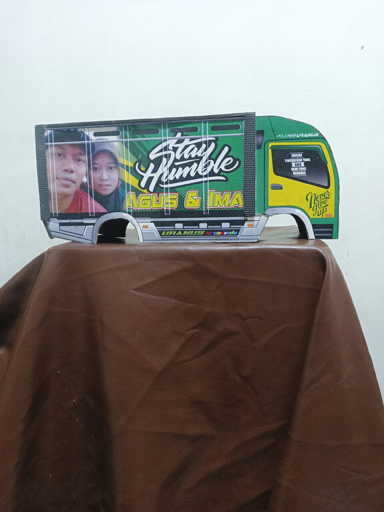 The image shows a miniature model of a green truck with yellow accents, placed on a brown, textured surface. The truck has a picture of a man and a woman on its side, with the text "Stay Humble AGUS & IMA" above them. On the driver's side door, there is text in Indonesian that reads "JANGAN TINGGALKAN YANG BAIK DEMI YANG MENARIK" which translates to "Don't leave the good for the attractive". Below this, it says "Never give up". The truck model is detailed, with simulated wheels and windows. The background is a plain white wall. The location within Penambangan, Indonesia is not specified.