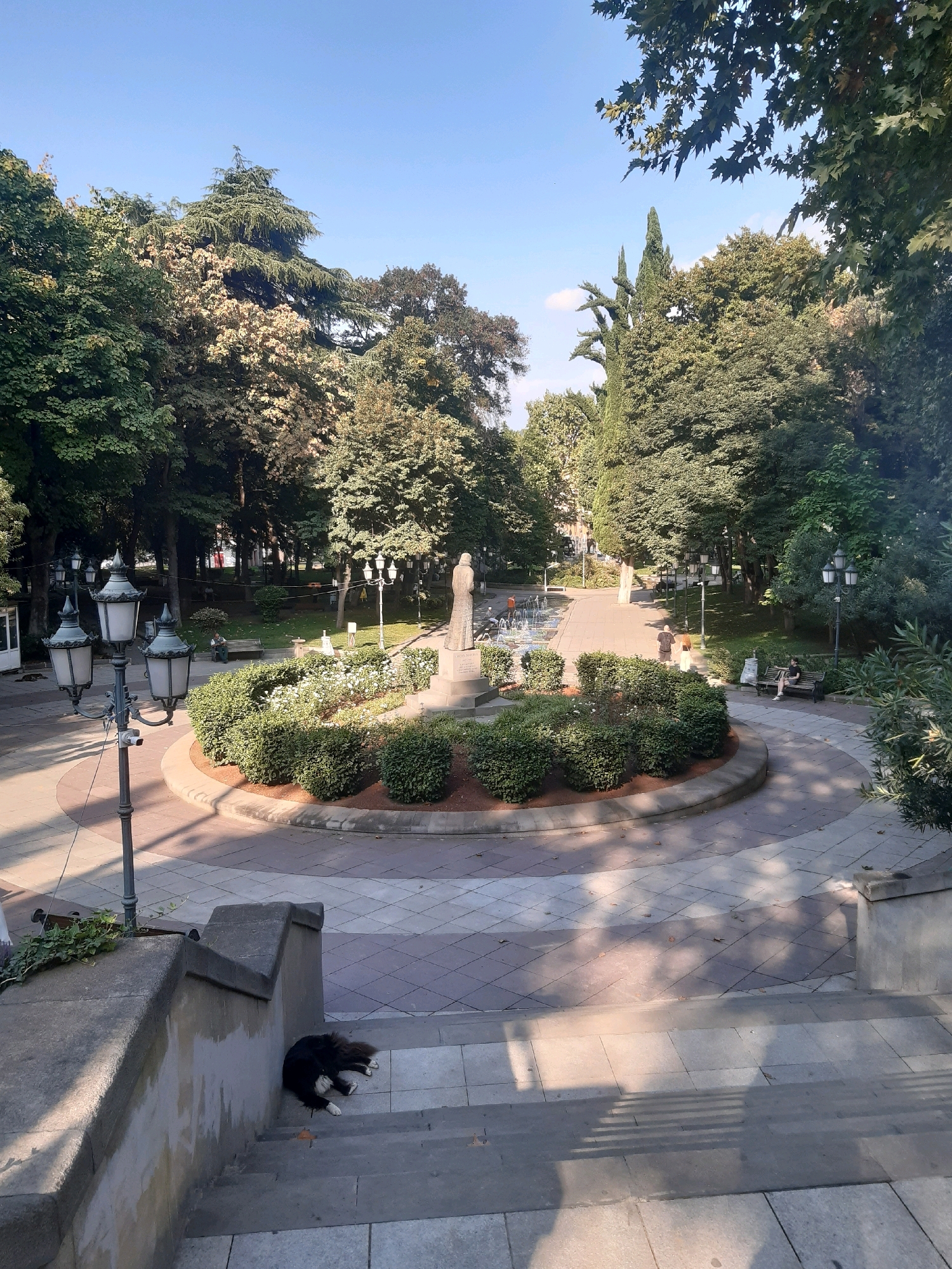 The image depicts a park scene in Tbilisi, Georgia. In the foreground, a black and white dog is sleeping on concrete steps. A lamppost with three lamps stands to the left. In the center, a circular flowerbed with white flowers and manicured bushes surrounds a statue of a person. A fountain with water jets is visible in the background, along with several people seated on benches and walking. The park is surrounded by lush green trees and a bright blue sky.