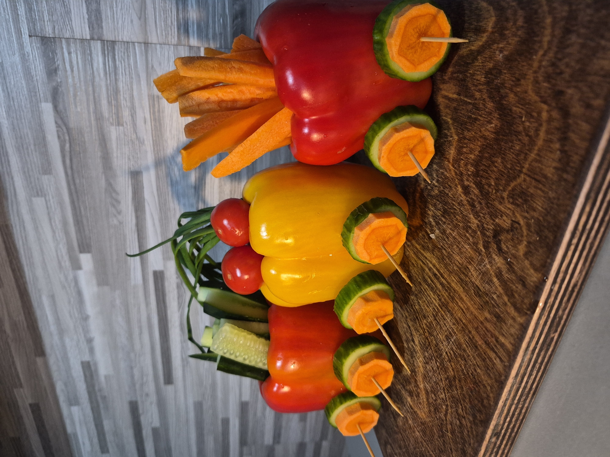 The image shows three bell peppers stacked vertically on top of each other, arranged to resemble a train or vehicle. The top pepper is red, the middle pepper is yellow, and the bottom pepper is orange. The peppers are "decorated" with other vegetables. The red pepper has orange carrot sticks sticking out of one end, resembling exhaust pipes, and sliced cucumbers and carrots are skewered to the sides with toothpicks to appear as wheels. The yellow pepper has cherry tomatoes and green onions sticking out of one end. The orange pepper has slices of cucumber sticking out of one end. The peppers are resting on a brown wooden surface that has a striped edge. Part of a light gray and white surface is visible beneath the wooden surface. The location is unknown.