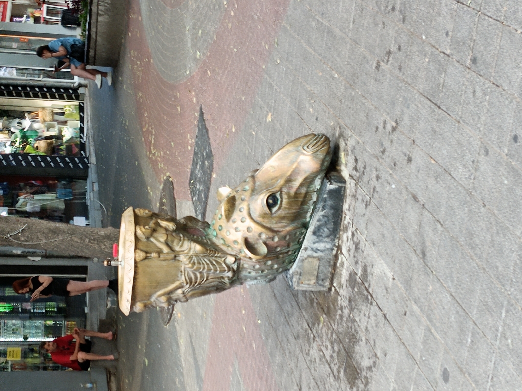 The image shows a street scene in Plovdiv, Bulgaria. In the foreground, a decorative fountain shaped like a fish head is visible, made of a weathered bronze-like material. A woman in a black dress and shorts is standing to the left of the fountain, looking at her phone. Further to the left, a man in a red shirt and dark shorts is sitting on the ground, also looking down. In the background, a person in a blue shirt and black shorts is leaning over, interacting with something out of frame, near a shop display. The setting appears to be a commercial area with shops and a sidewalk. The lighting suggests it is daytime. The overall atmosphere is casual, with people engaged in their own activities. There are no visible signs with text in this image.