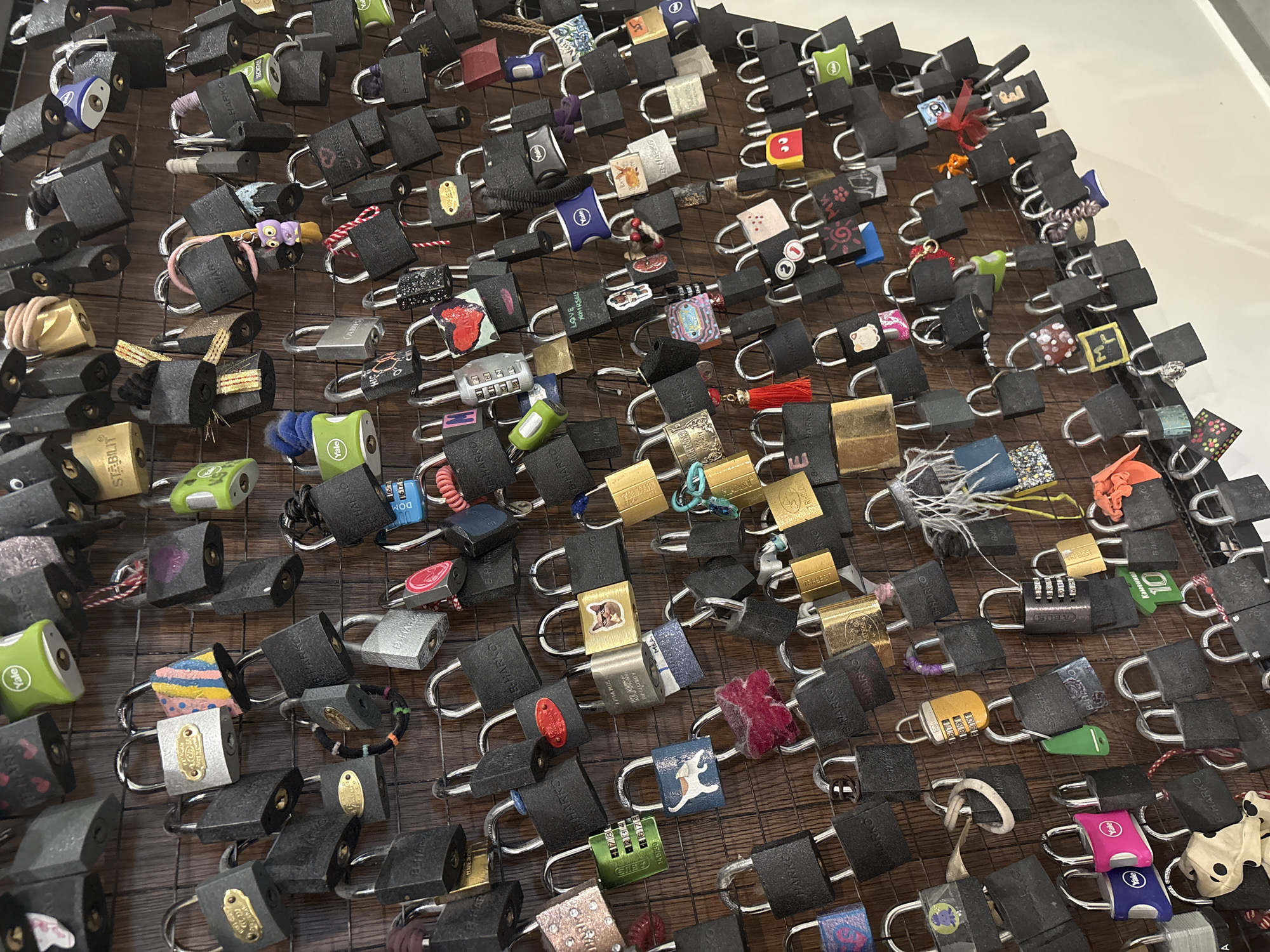 The image shows a close-up view of a dense collection of padlocks attached to a wire grid. The padlocks are of various sizes, colors, and designs, indicating that they have been left by different individuals. Many of the padlocks have black bodies, while the hasps are typically silver or chrome. Some padlocks have unique decorations, such as stickers, painted designs, or small trinkets attached to them. This suggests the padlocks are 'love locks' symbolizing commitment or affection. The location is Unknown, Unknown.
