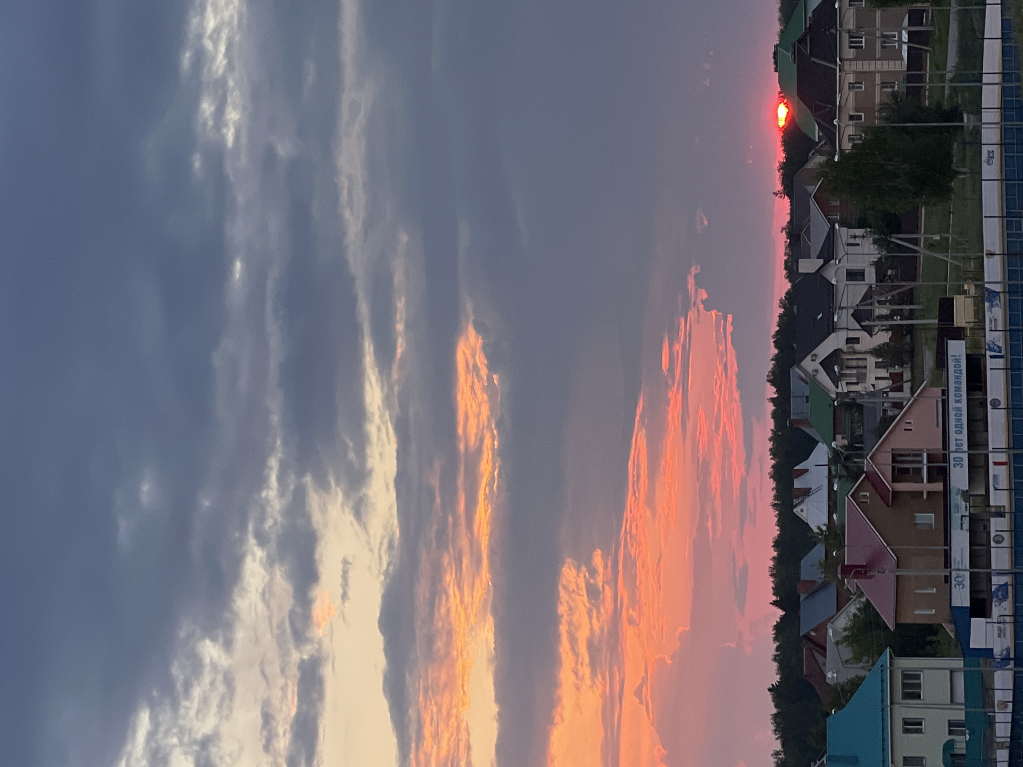 The image depicts a sunset over a residential area in Noviy, Russia. The sky is filled with dramatic clouds, transitioning from pale yellow near the horizon to deep gray overhead. The setting sun, a vibrant red orb, is partially obscured by trees and the roofs of houses. The houses themselves are of varying architectural styles, with many featuring pitched roofs. Some of the houses are illuminated by the fading sunlight, while others are cast in shadow. In the foreground, there appears to be a fence or barrier, possibly part of a sports facility, with some signage visible. The overall impression is of a serene, yet visually striking, end to the day.