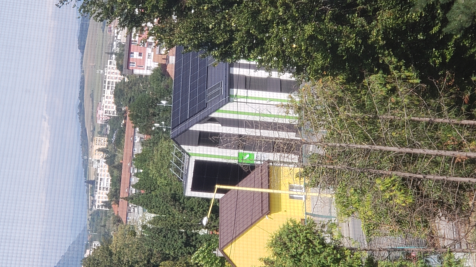 The image depicts a residential area in Prešov, Slovakia, featuring a blend of modern and older structures, as well as abundant greenery. In the foreground, a bright yellow house with a brown roof stands adjacent to a more contemporary building. This modern structure is characterized by its white facade with vertical green stripes, a sloping dark roof fitted with solar panels, and large dark windows. Several trees, some barren and others lush green, are scattered throughout the scene, with a prominent evergreen tree on the right side. In the background, a cityscape of multi-story buildings with red and white facades is visible, nestled against a backdrop of rolling hills and a hazy sky. The overall impression is one of a tranquil, suburban environment with a mix of residential development and natural landscapes.