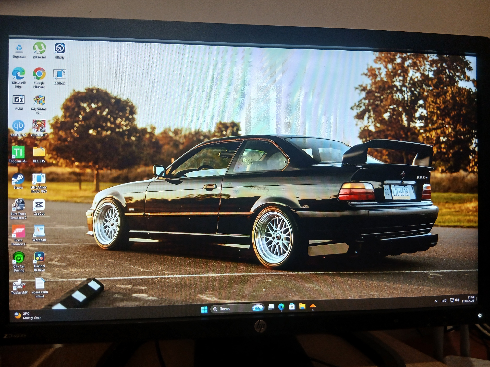 The image shows a computer monitor displaying a desktop background of a black BMW M3 coupe. The car is parked on asphalt, with trees and a bright sky in the background. The desktop icons are arranged in a grid on the left side of the screen. The taskbar at the bottom displays the time, date, network status, and other system icons. The monitor is placed on a wooden surface. There are no people or animals visible in the image. The scene is well-lit and the image is clear. The specific location in the city of Dnipro, Ukraine, cannot be determined from the image alone.