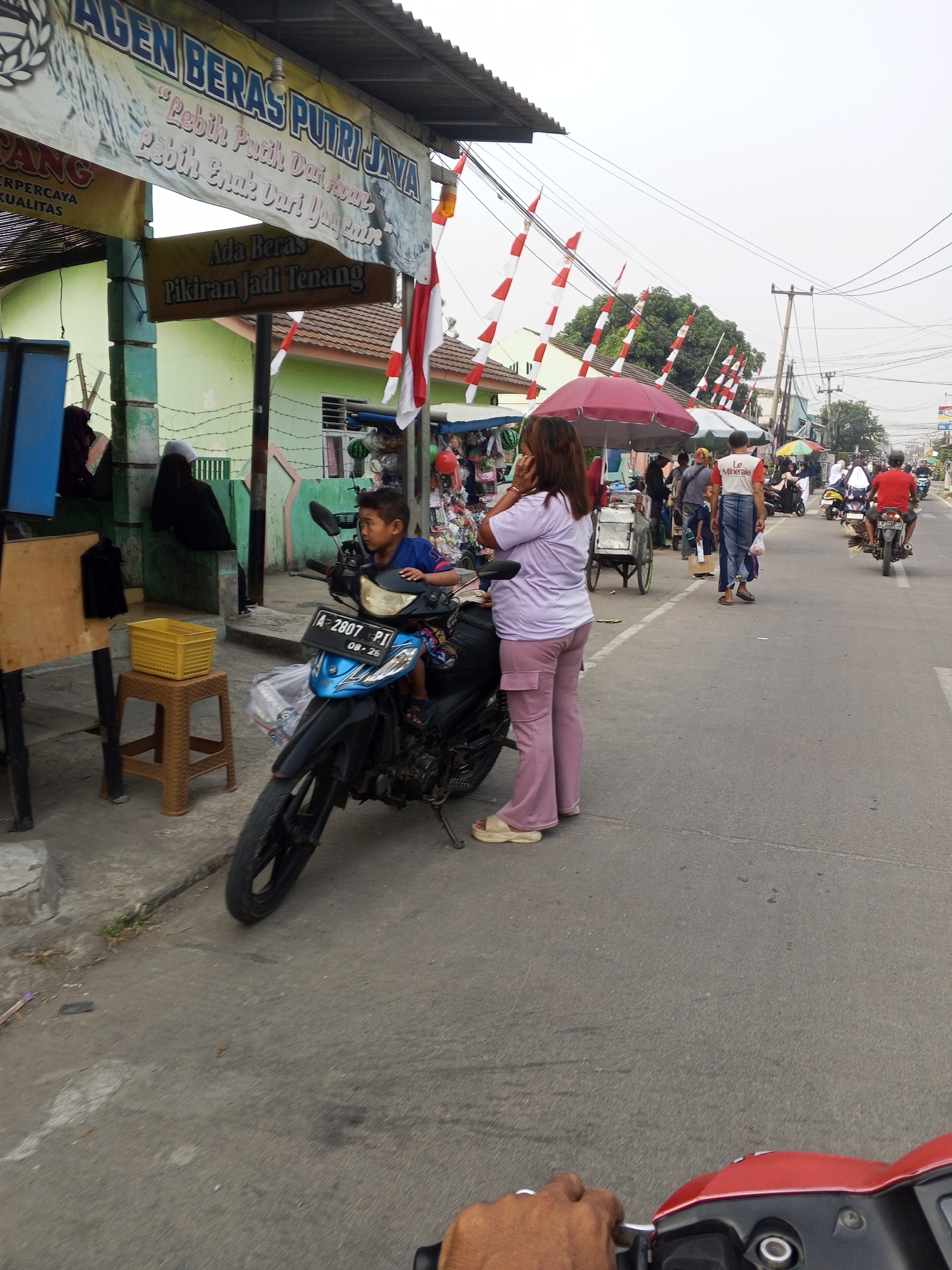 The image depicts a street scene in Ciruas, Indonesia, with a focus on a motorcycle and its young rider. The rider, a boy wearing a blue shirt, is seated on a blue and black motorcycle. To his left, a woman in a light purple shirt and pink pants stands near the motorcycle. Behind her, a sign reads "Agen Beras Putri Jaya" with Indonesian text below. Further in the background, there are market stalls and several people. Red and white Indonesian flags are visible on poles. The street is paved, and there are buildings and trees lining the road. The sky is overcast.
